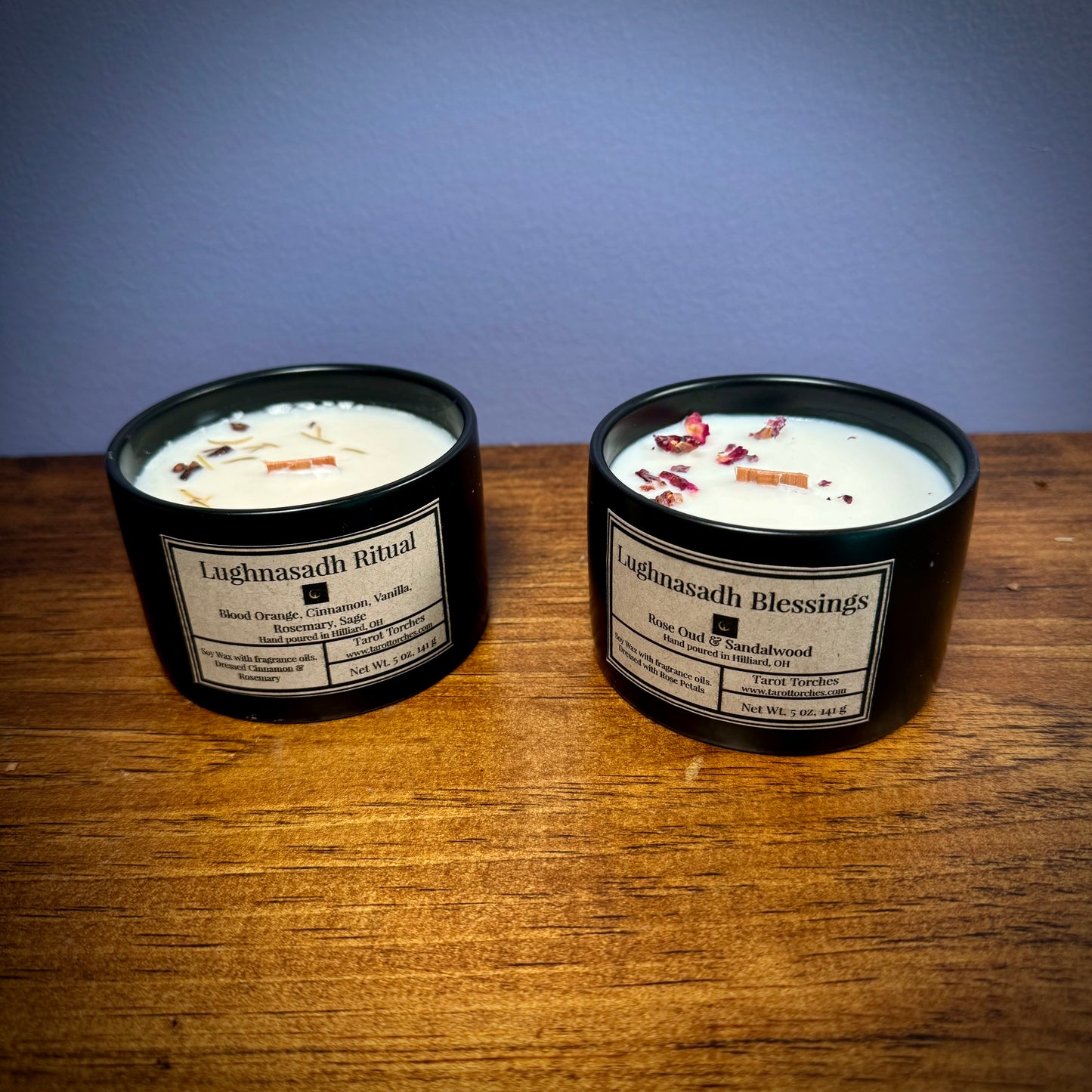 Two ceramic candles with pine lids, labeled 'Lughnasadh Blessings' and 'Lughnasadh Ritual', placed on a wooden surface against a purple background.
