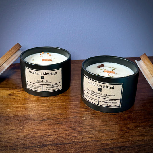 Two ceramic candles on a wooden surface, one labeled 'Samhain Blessings' and the other 'Samhain Ritual', with visible wax and natural herb toppings.