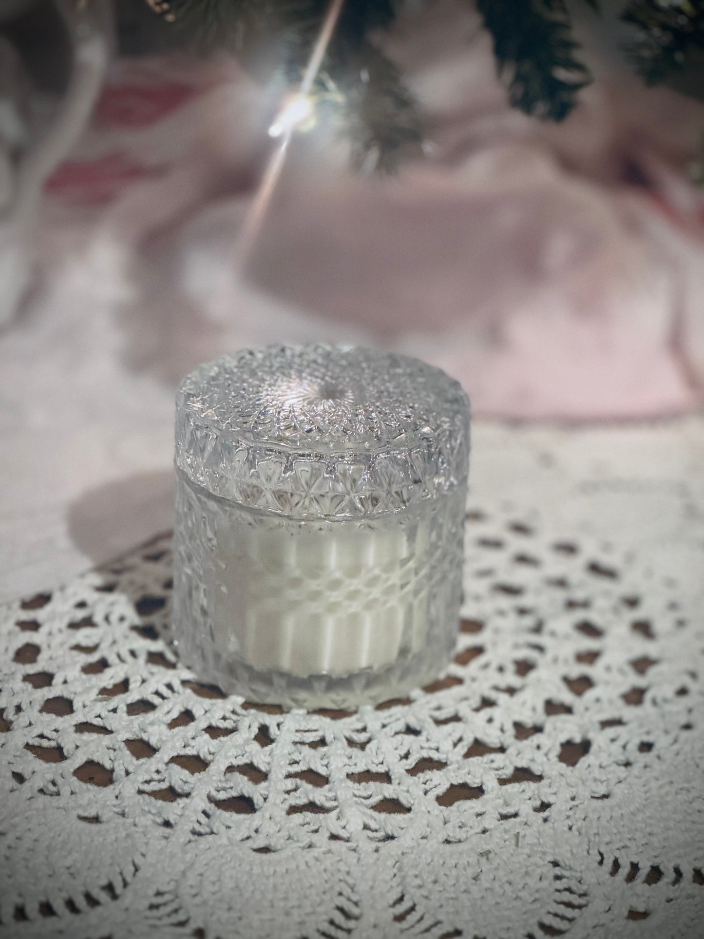 A clear glass candle vessel with lid placed on a lace tablecloth in front of a decorated Christmas tree.