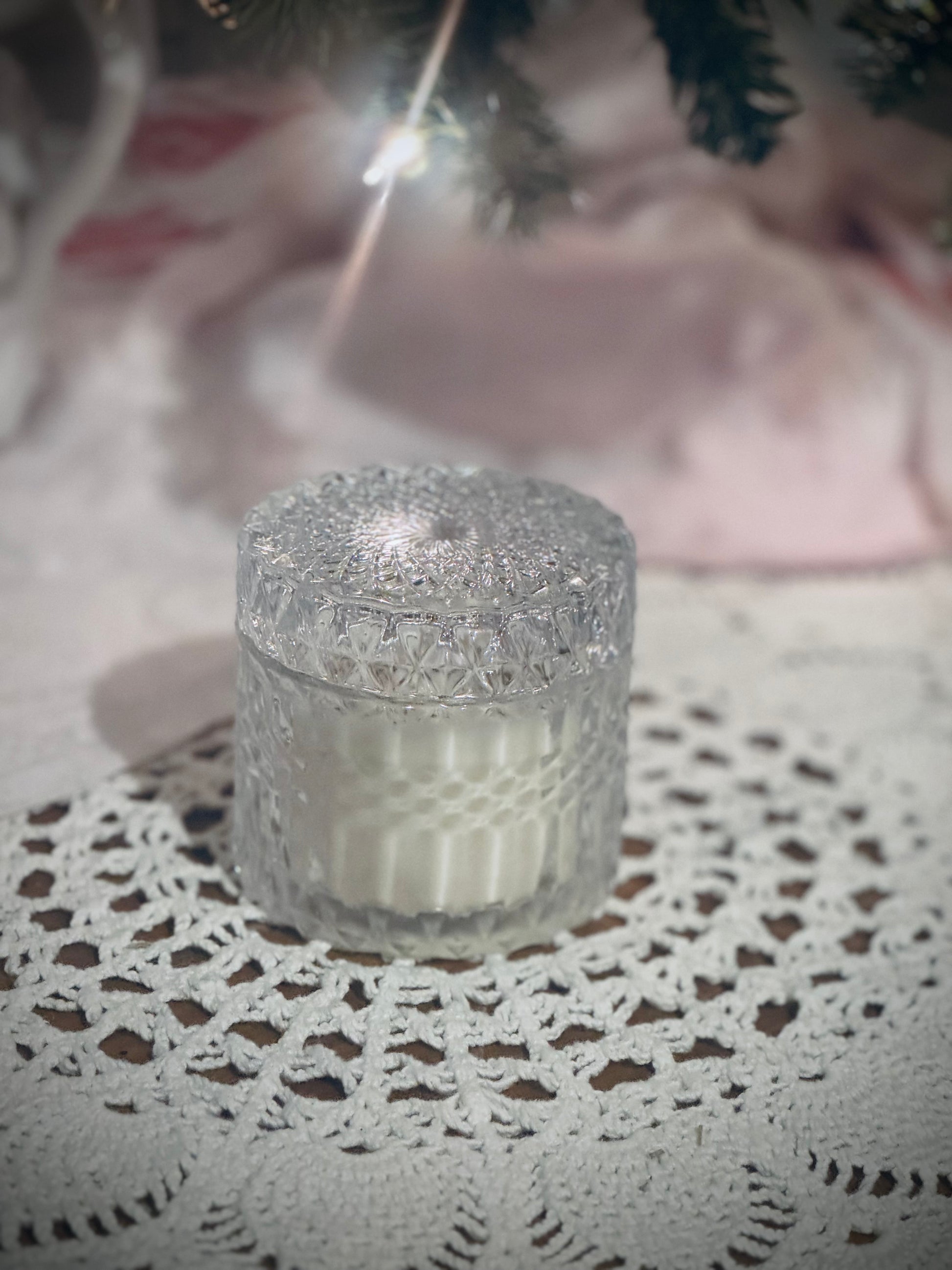 A clear glass candle vessel with lid placed on a lace tablecloth in front of a decorated Christmas tree.