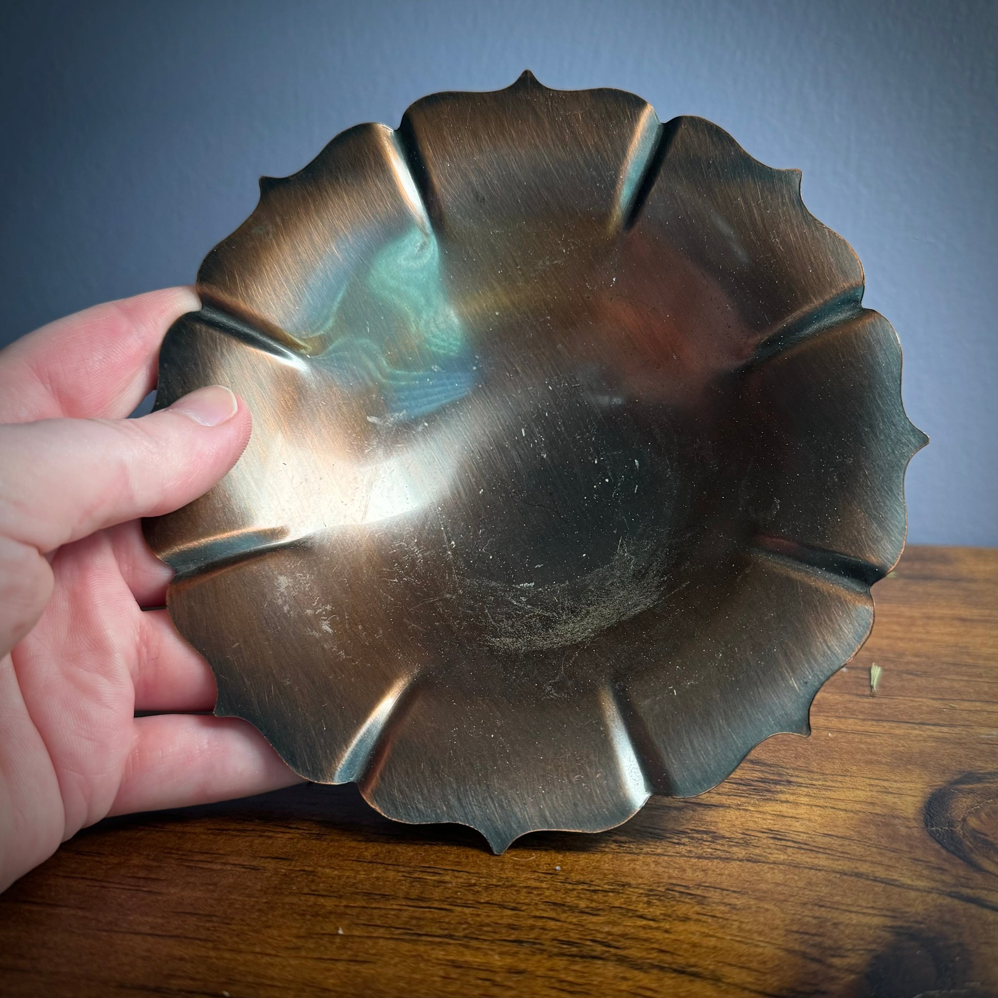 A vintage ritual bowl with a scalloped edge and a deep dark copper color, being held over a wood surface.