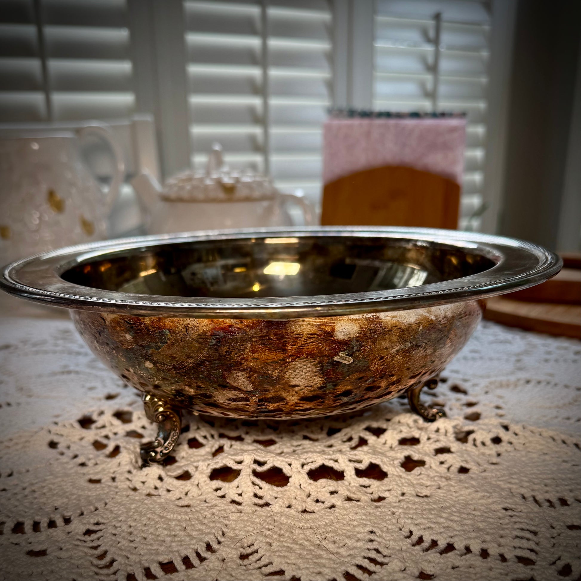 A vintage silverplate footed altar bowl is displayed on a table with a lace tablecloth, positioned in front of a window with natural light coming through.