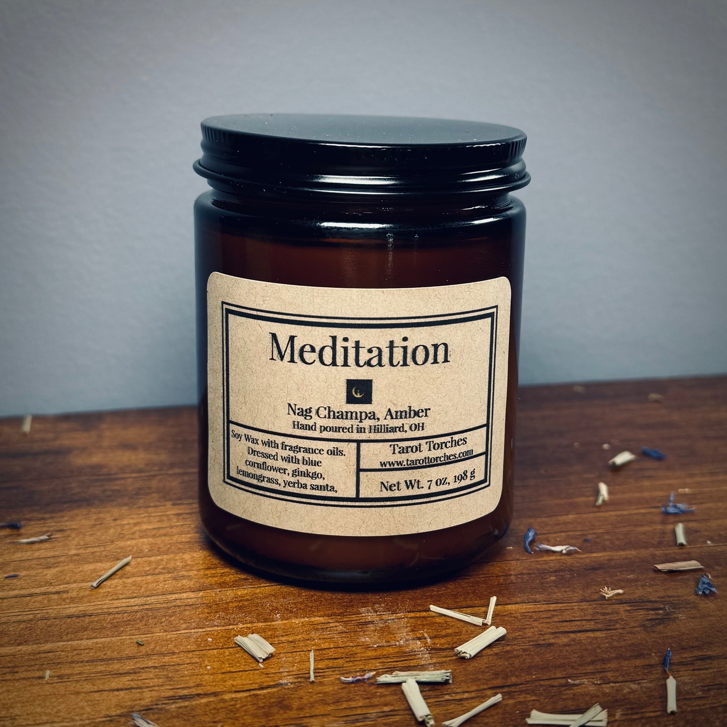 An amber glass candle reading "Meditation" sits on a wooden surface with scattered herbs around.