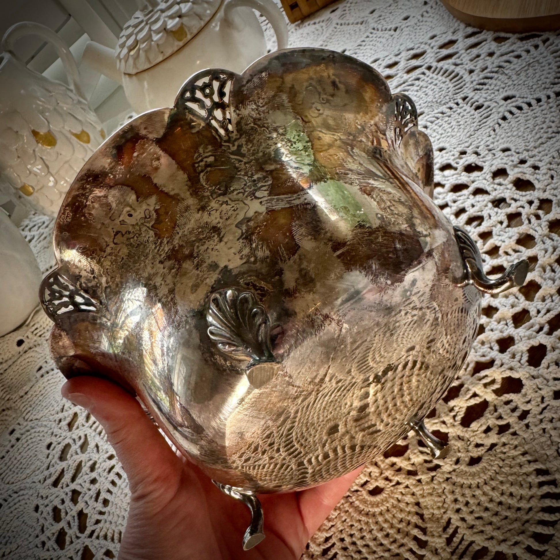 A vintage silver serving bowl with scalloped edges and feet, placed on a table with a lace tablecloth.