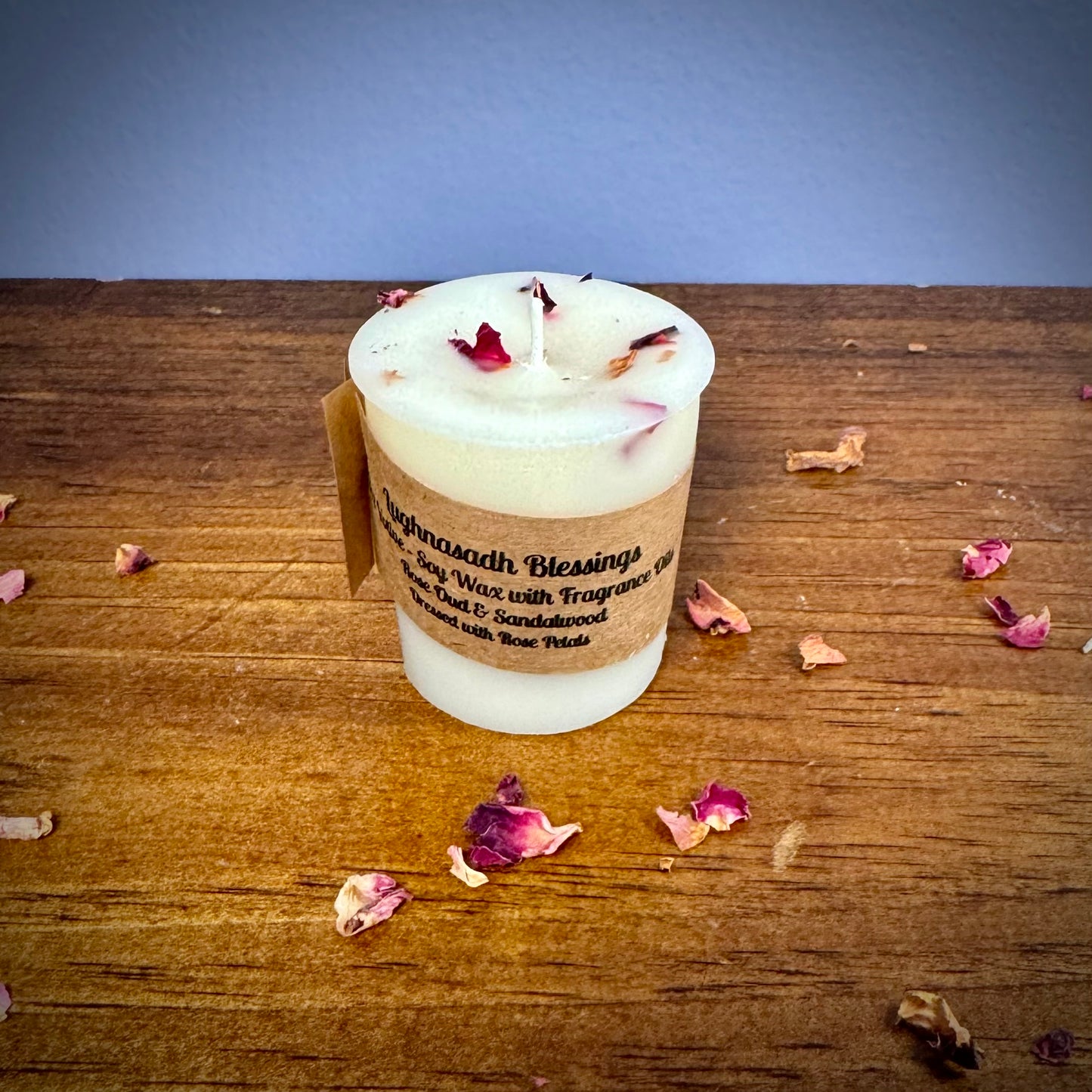 A white votive candle with a label reading "Lughnasadh Blessings" on a wooden surface.