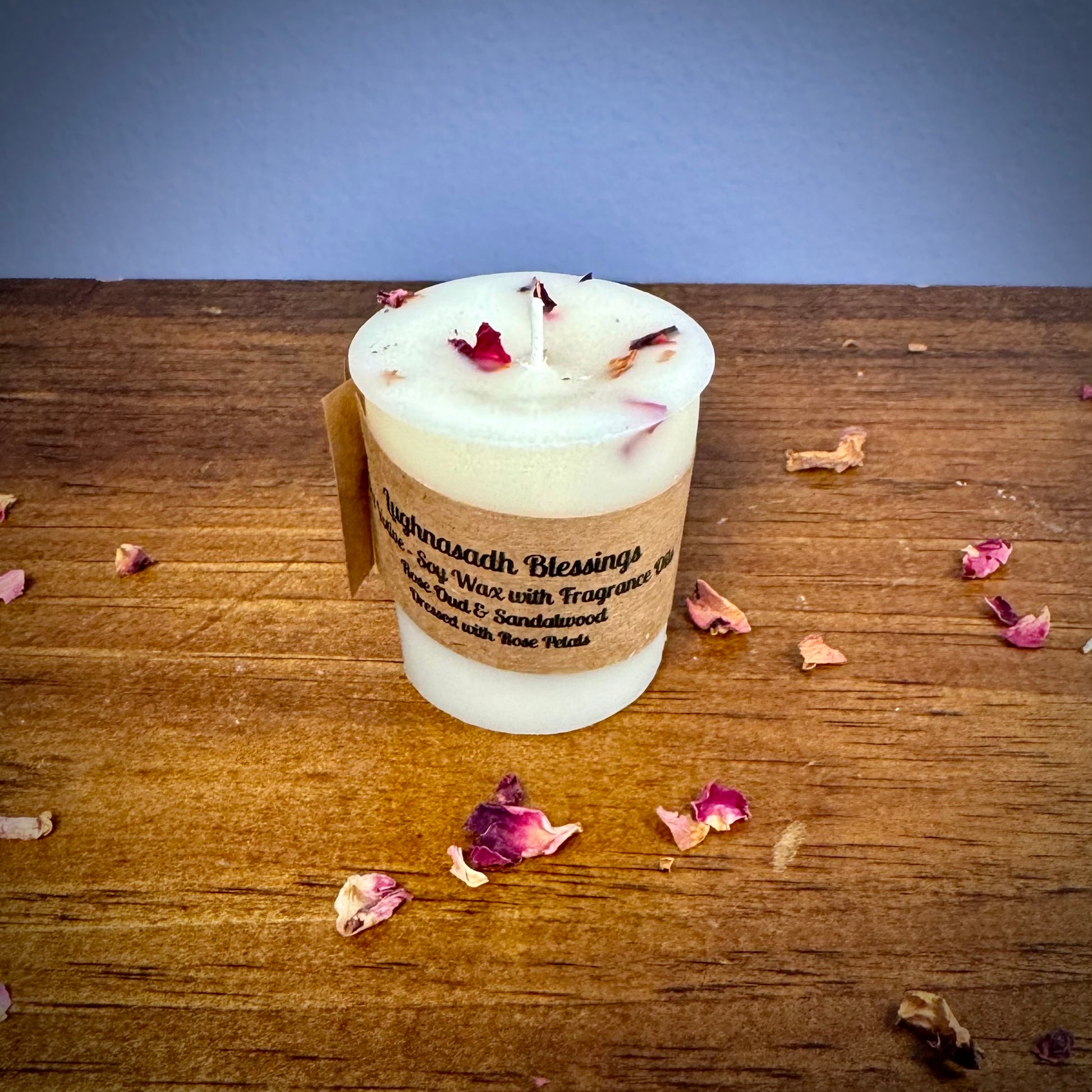 A white votive candle with a label reading "Lughnasadh Blessings" on a wooden surface.