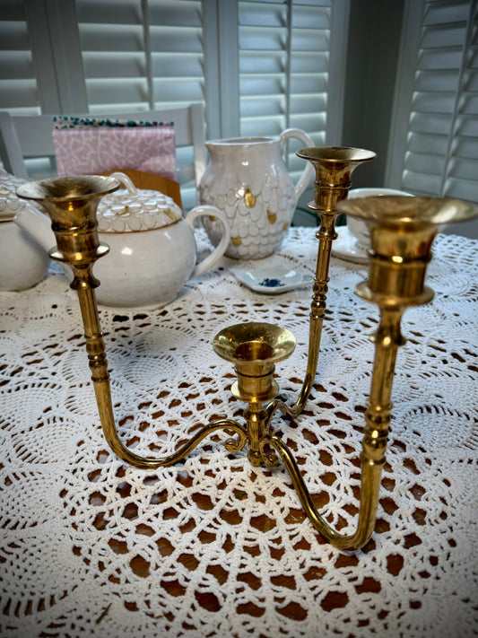 A vintage brass four-arm candelabra placed on a table with a lace tablecloth, with pottery in the background.
