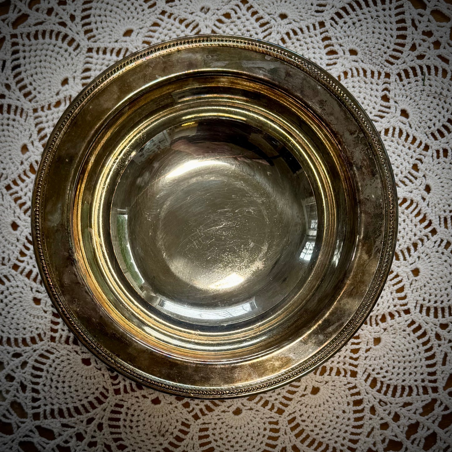 A vintage silverplate footed altar bowl is displayed on a table with a lace tablecloth, positioned in front of a window with natural light coming through.