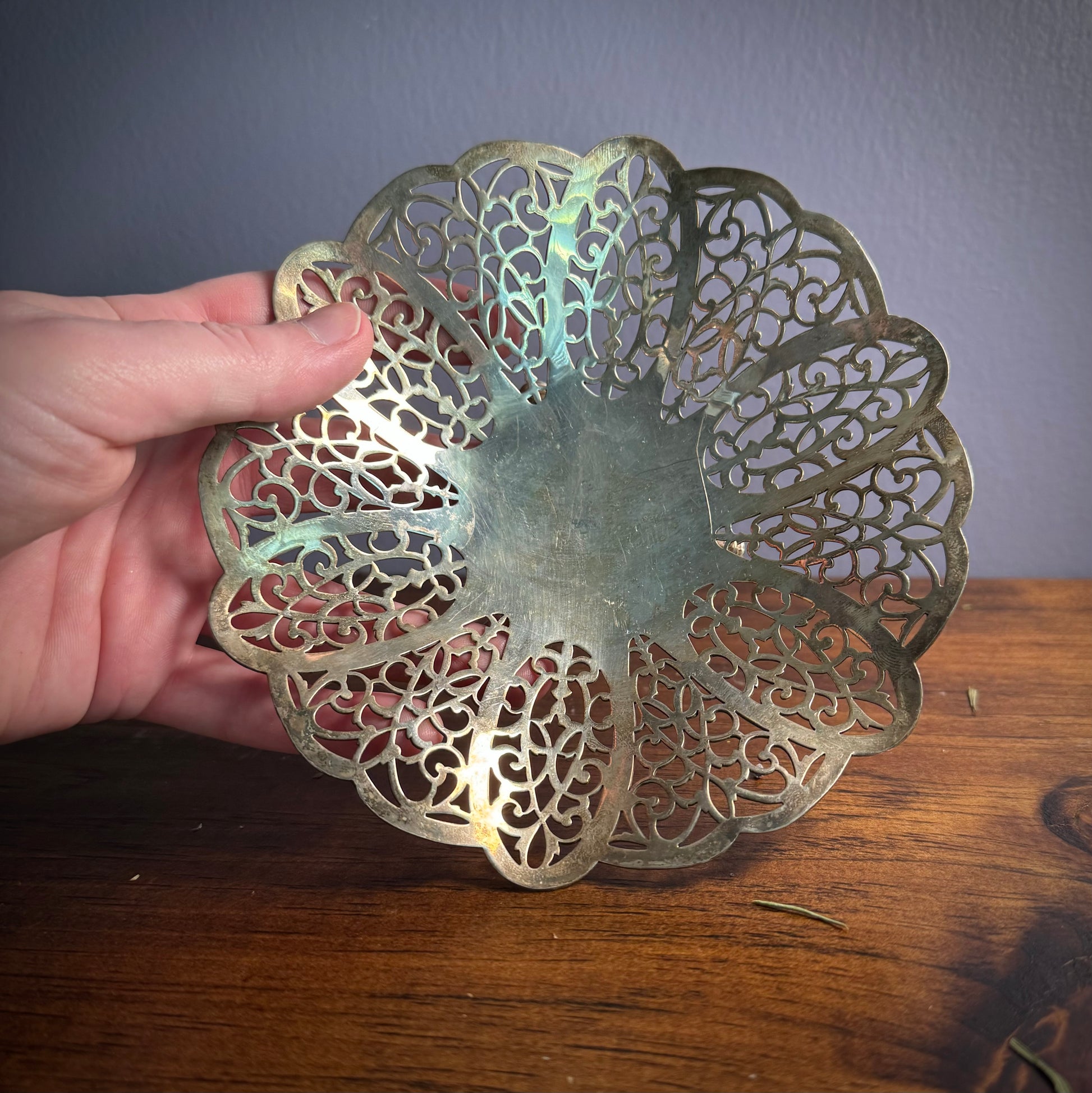A vintage silver ritual plate with intricate floral lace patterns being held over a wood surface.