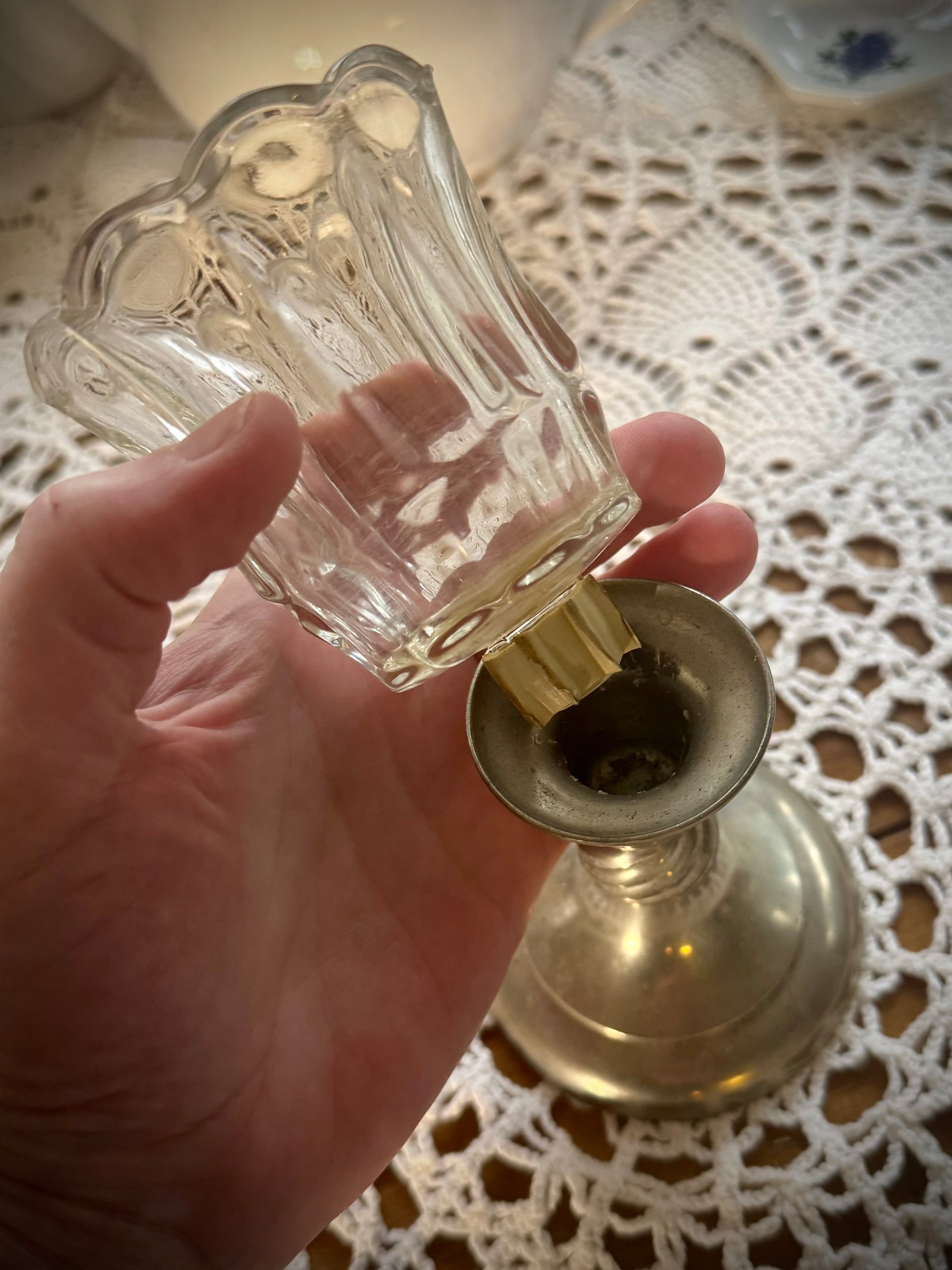 A clear glass votive candle holder with a textured design and a wooden peg at the bottom, placed on a lace tablecloth.