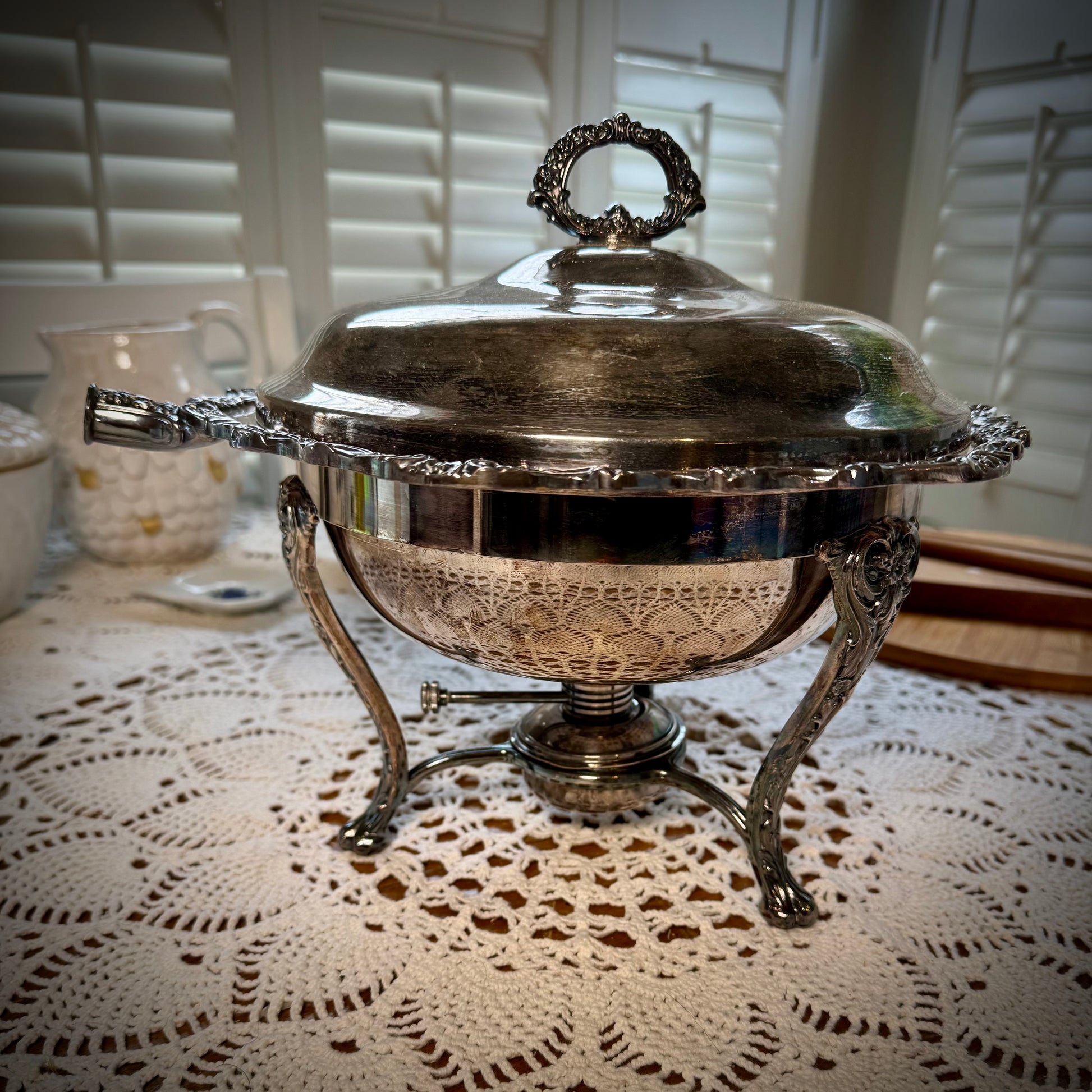 A vintage silver chafing dish with a lid, handle detached, placed on a table with a lace tablecloth.