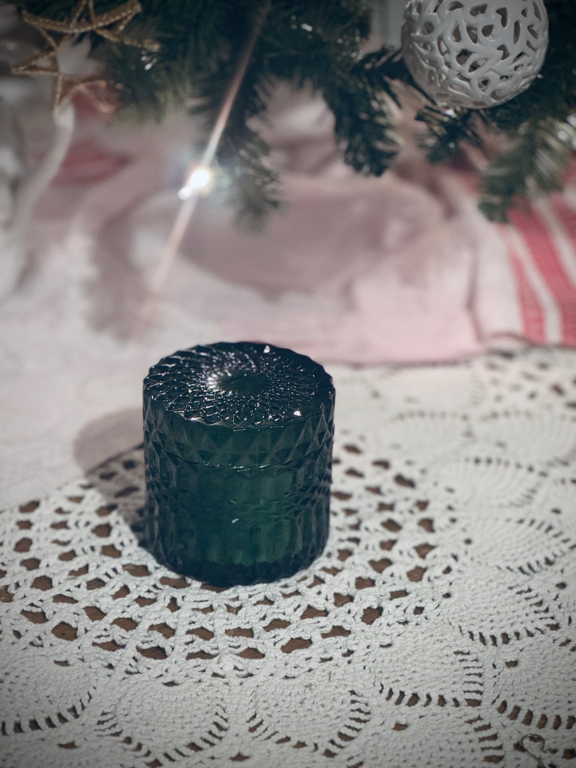 A green glass candle vessel with lid placed on a lace tablecloth in front of a decorated Christmas tree.