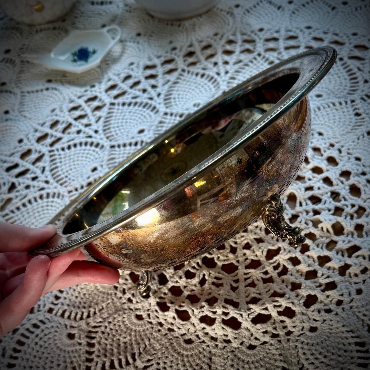 A vintage silverplate footed altar bowl is displayed on a table with a lace tablecloth, positioned in front of a window with natural light coming through.
