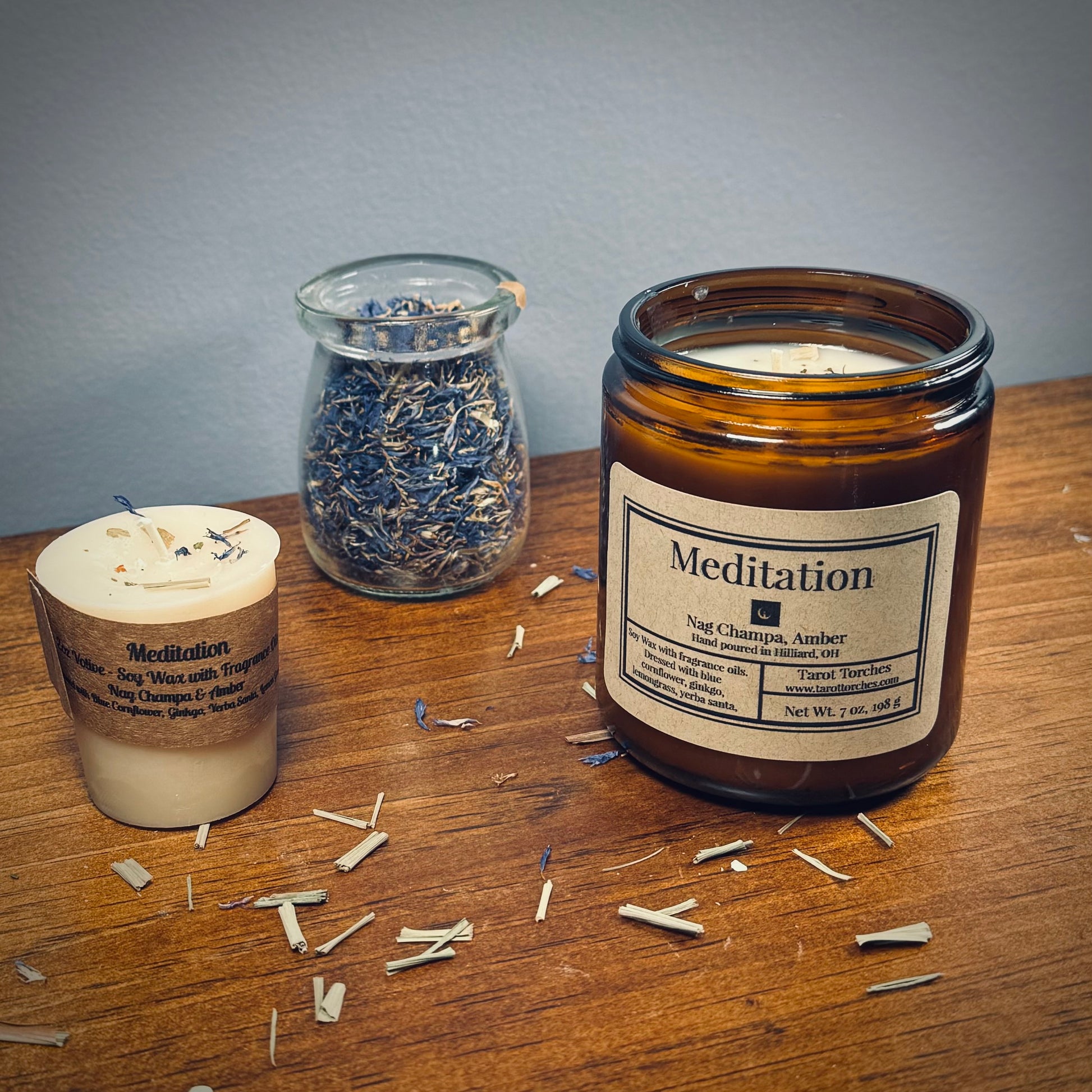 A white votive candle and amber glass candle both reading "Meditation" sit on a wooden surface with a glass jar of herbs between them.