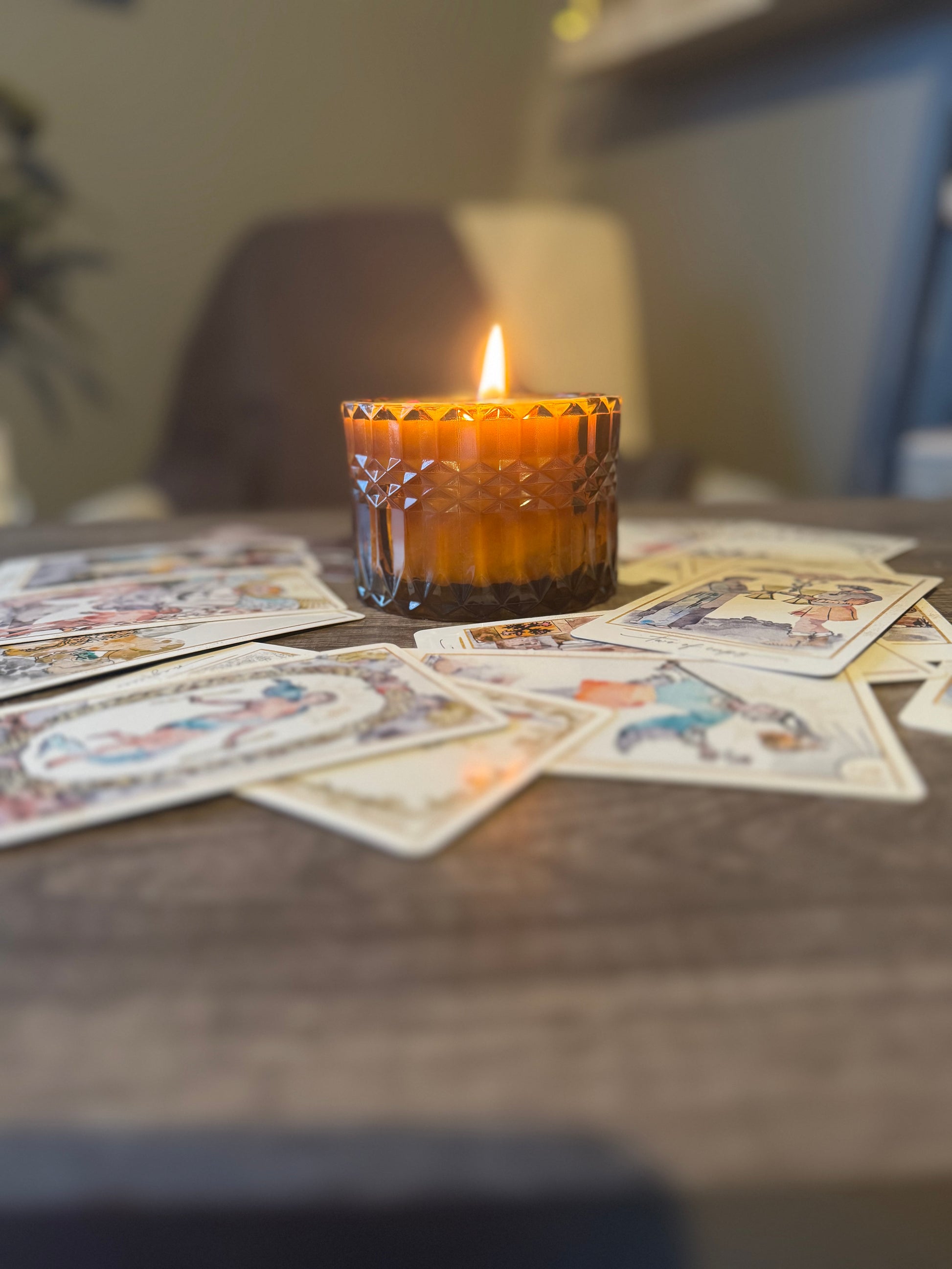 A soy wax candle with a cotton wick is burning in an amber glass jar with a lid, surrounded by various tarot cards spread out on a table.