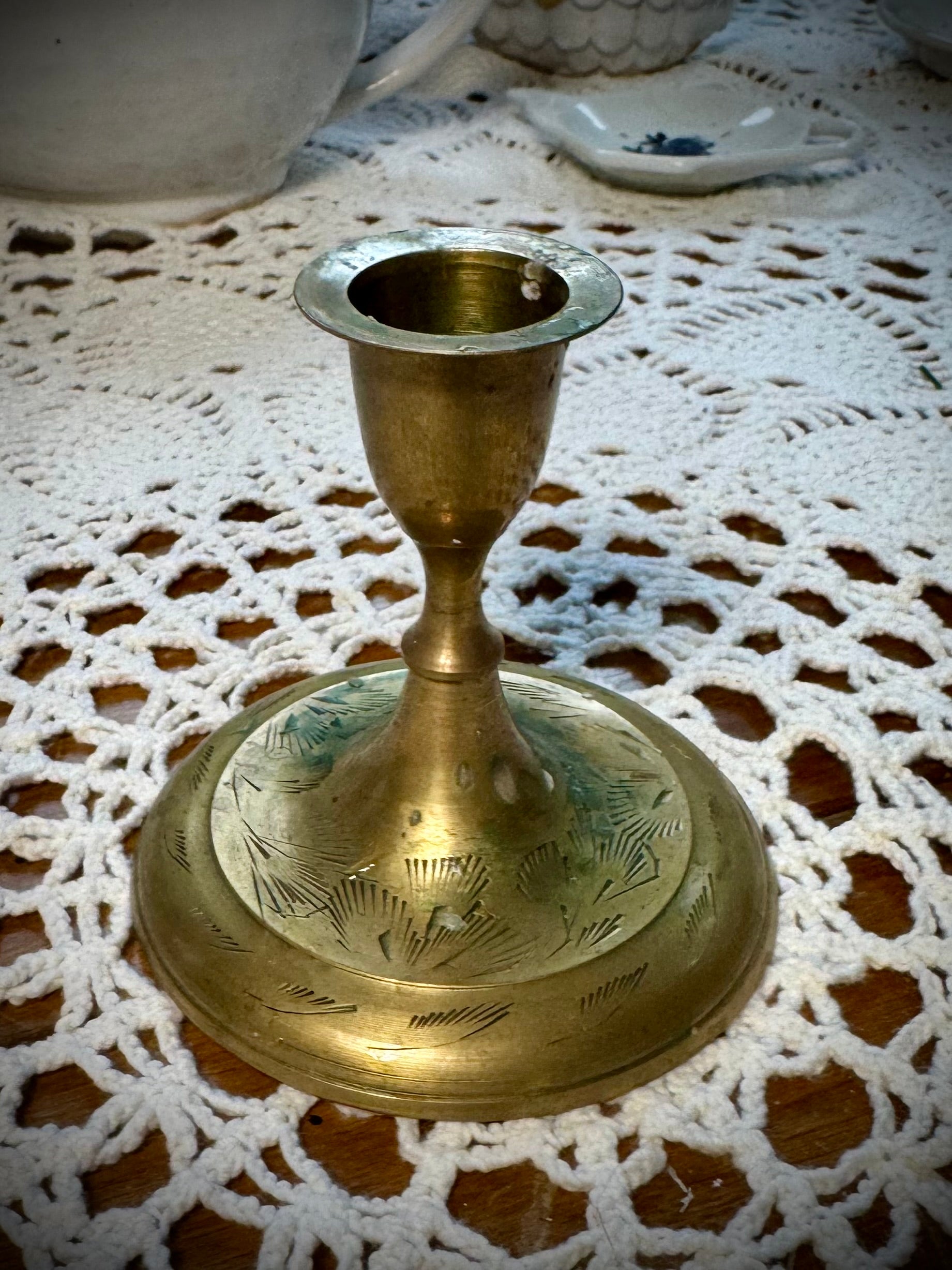 A vintage brass taper candle holder with etching on the base, displayed on a lace tablecloth.