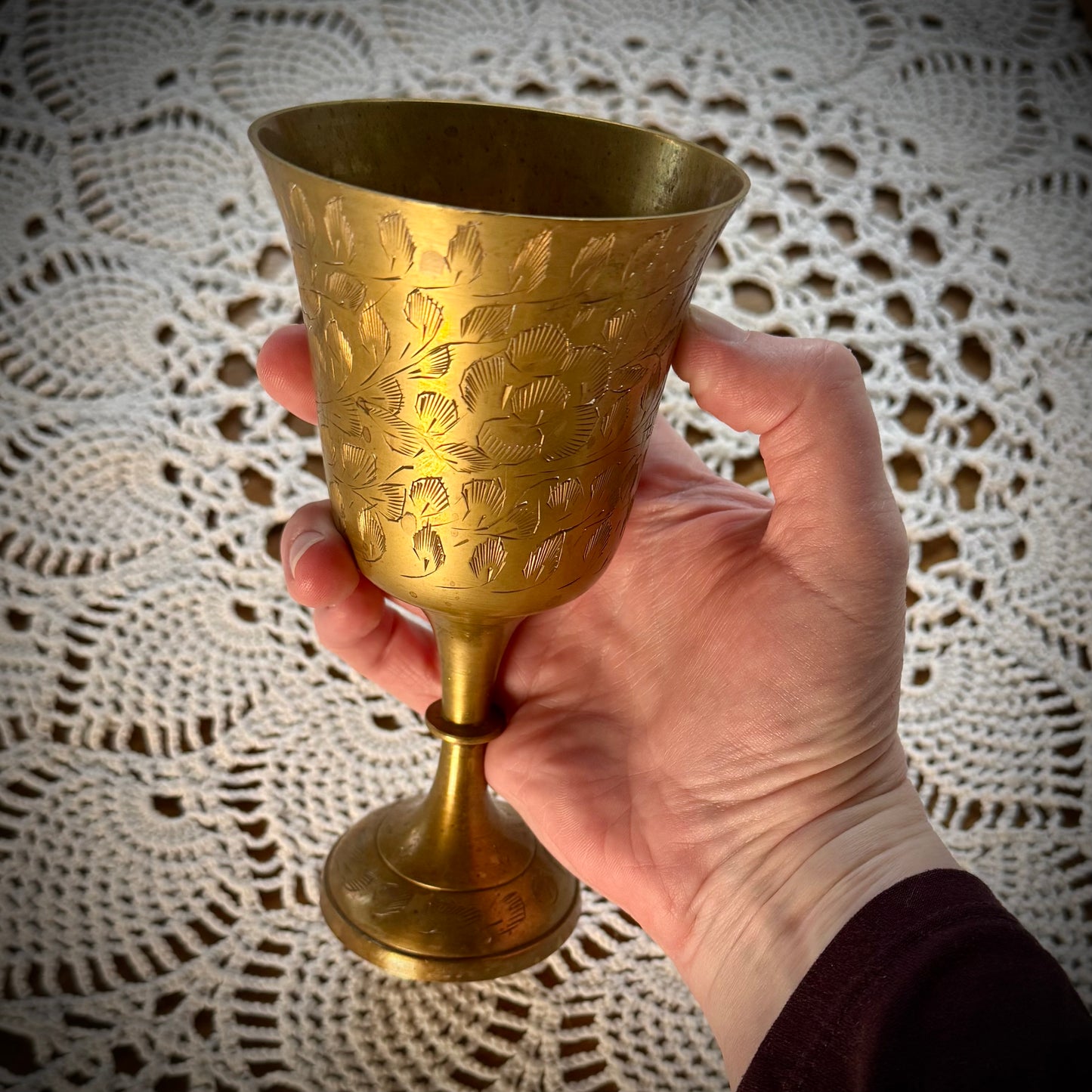 Etched brass colored goblet placed being held over a table with a white lace tablecloth.