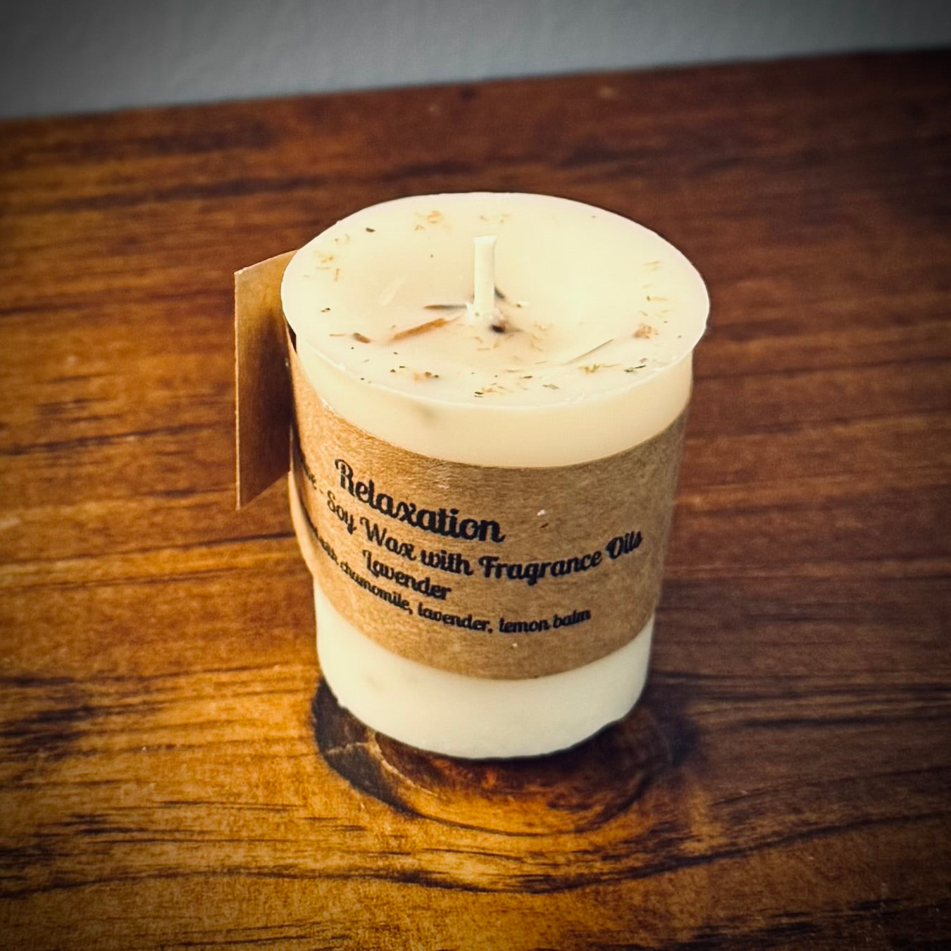 A soy wax votive candle with a cotton wick, labeled 'Relaxation' with natural herbs on a wooden surface.