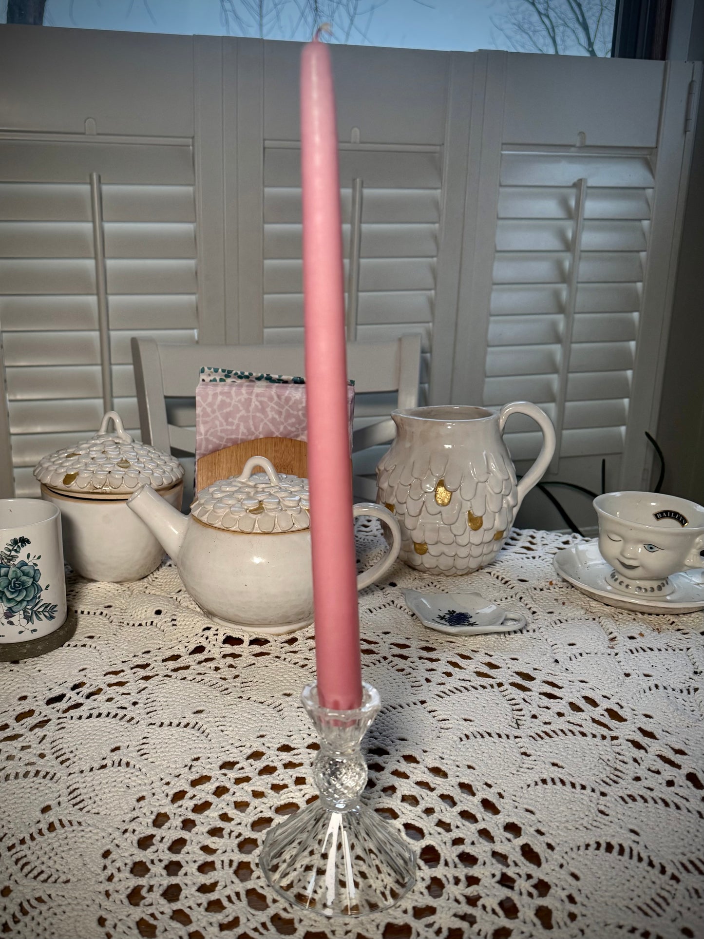 A small vintage clear glass candlestick on a table with a lace tablecloth holding a tall pink taper candle.