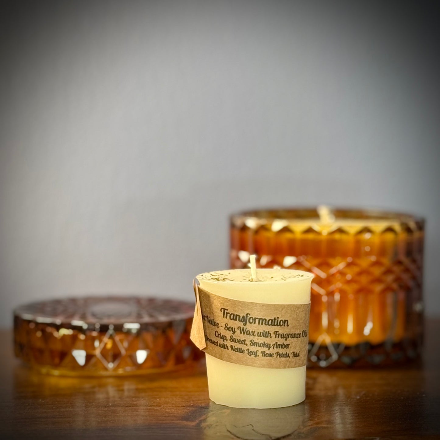 A small white votive with a kraft label sit on a table in front of an amber glass jar.