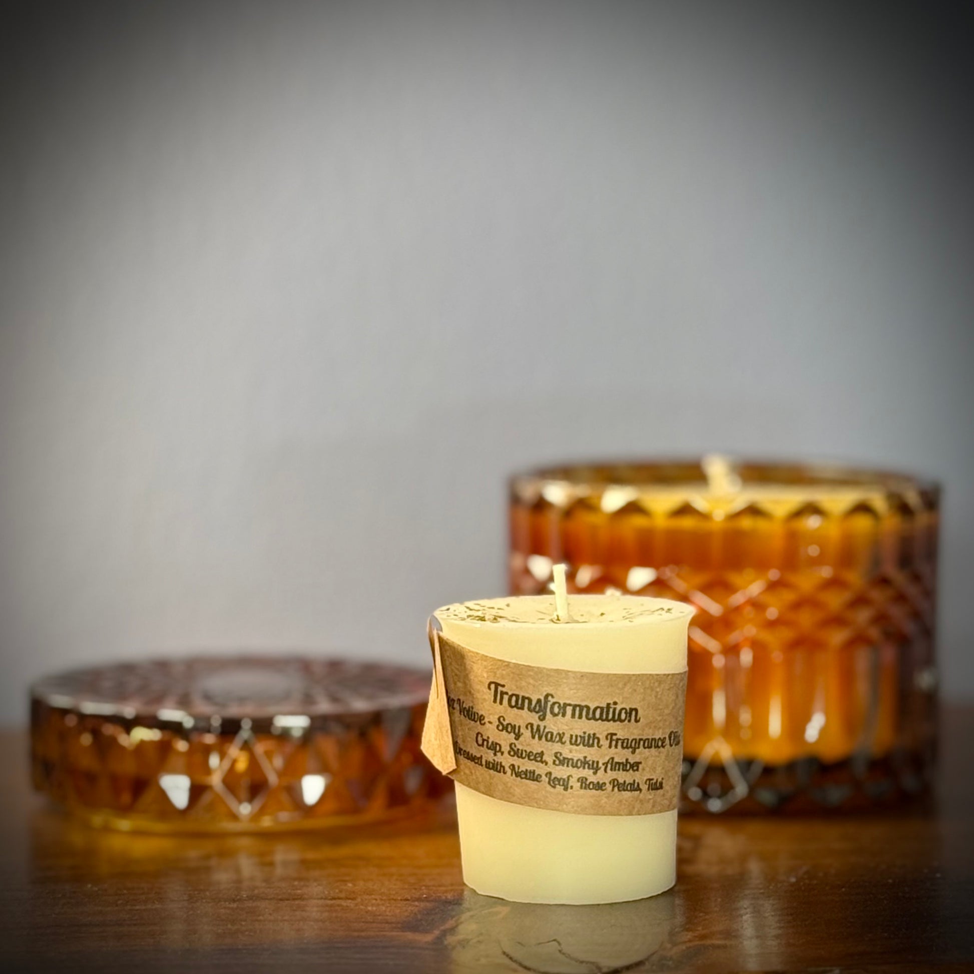 A small white votive with a kraft label sit on a table in front of an amber glass jar.