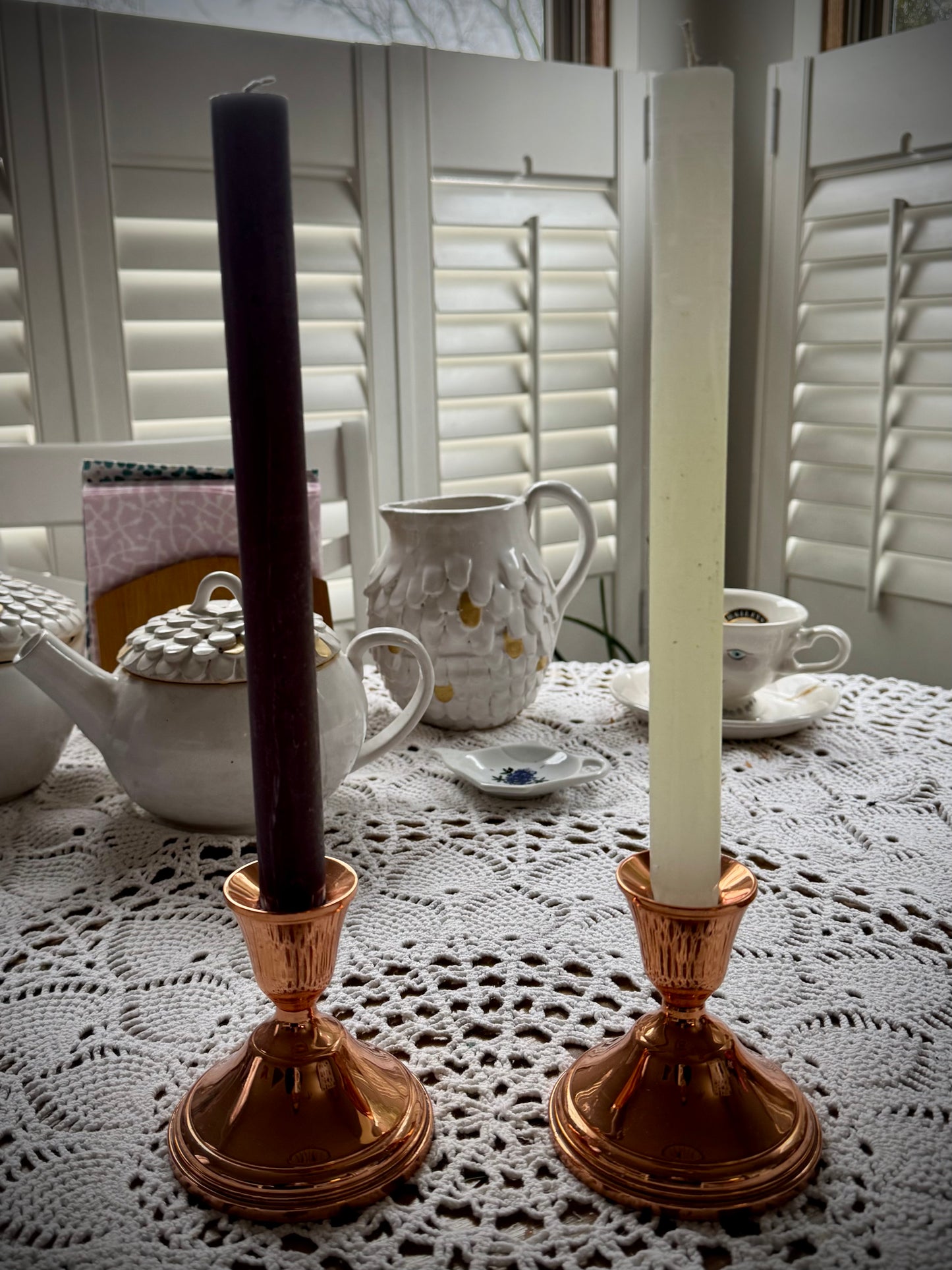 Two vintage copper candle holders displayed on a lace tablecloth, each holding a different color taper candle.