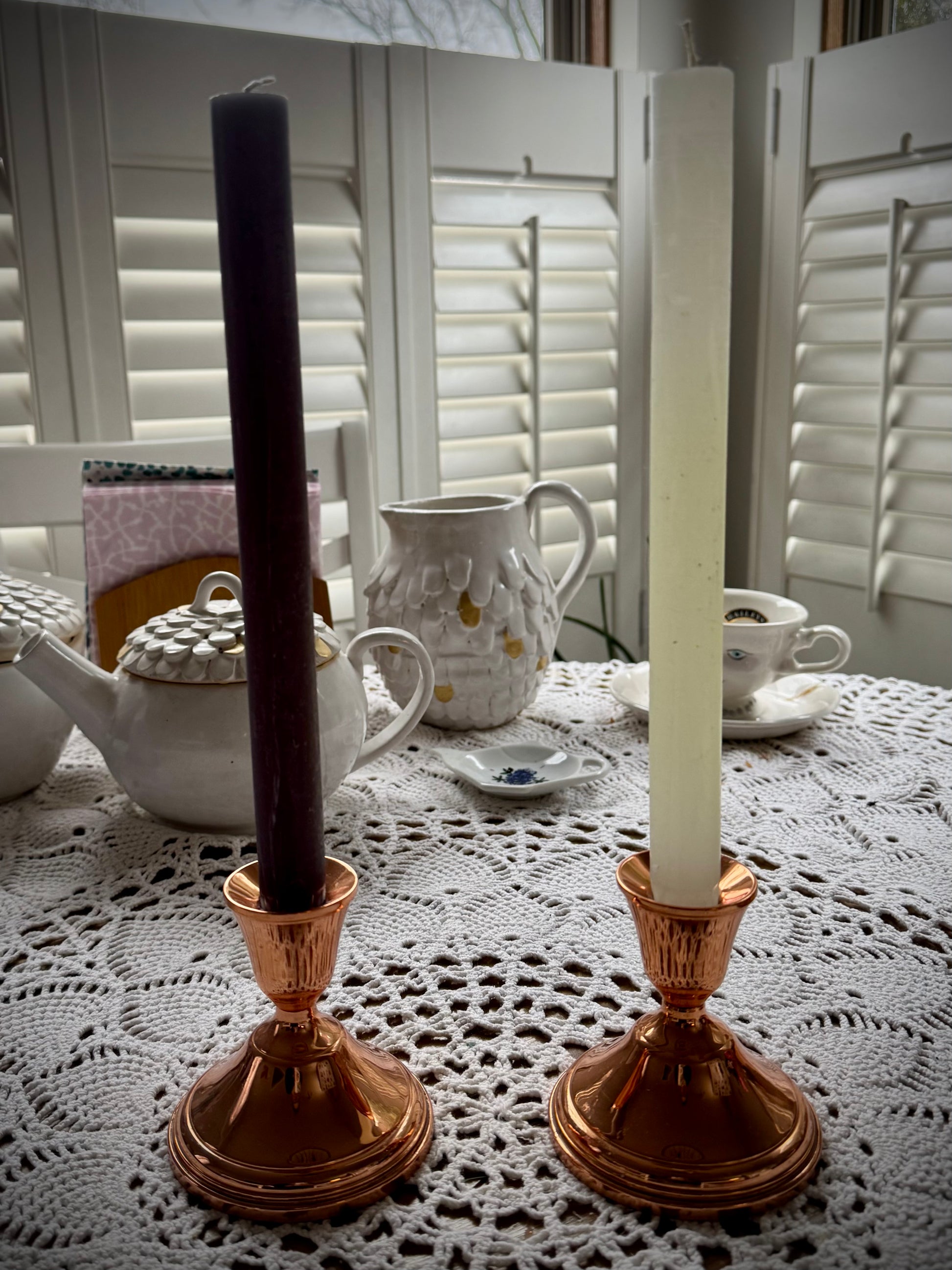 Two vintage copper candle holders displayed on a lace tablecloth, each holding a different color taper candle.