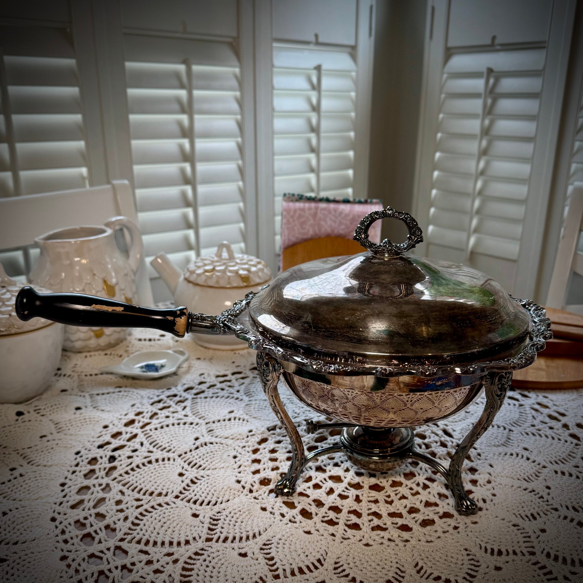 A vintage silver chafing dish with a lid, placed on a table with a lace tablecloth.