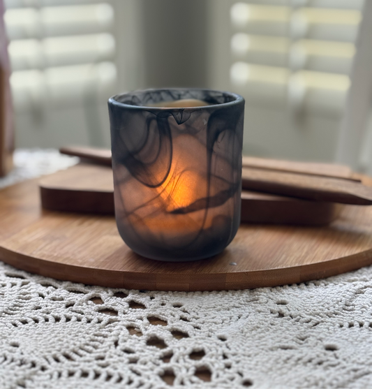 A soy wax candle with a black swirl vessel, lit and placed on a wooden surface with a lace tablecloth underneath.