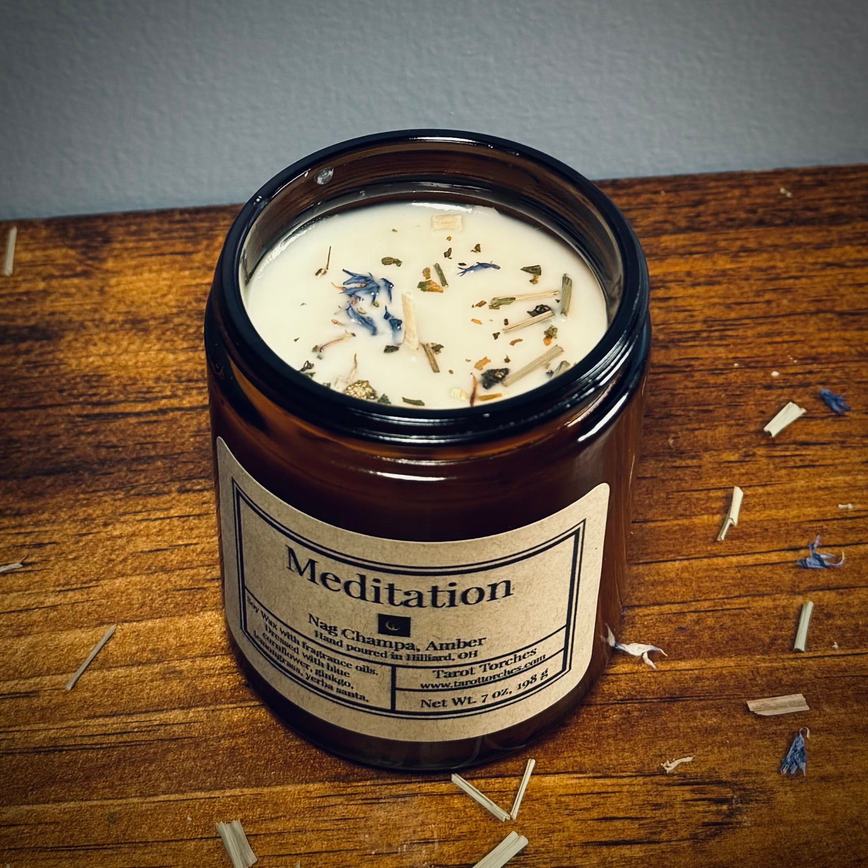 An amber glass candle reading "Meditation" sits on a wooden surface with scattered herbs around.