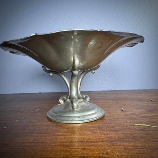A silver pewter bowl with three detailed feet attached to a round base, placed on a wooden surface with a plain blue background.