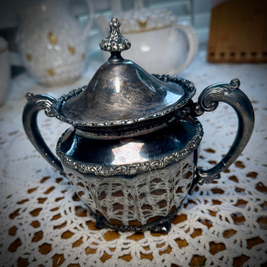 Vintage silver plate sugar bowl with intricate designs, showing signs of wear, placed on a white lace tablecloth.