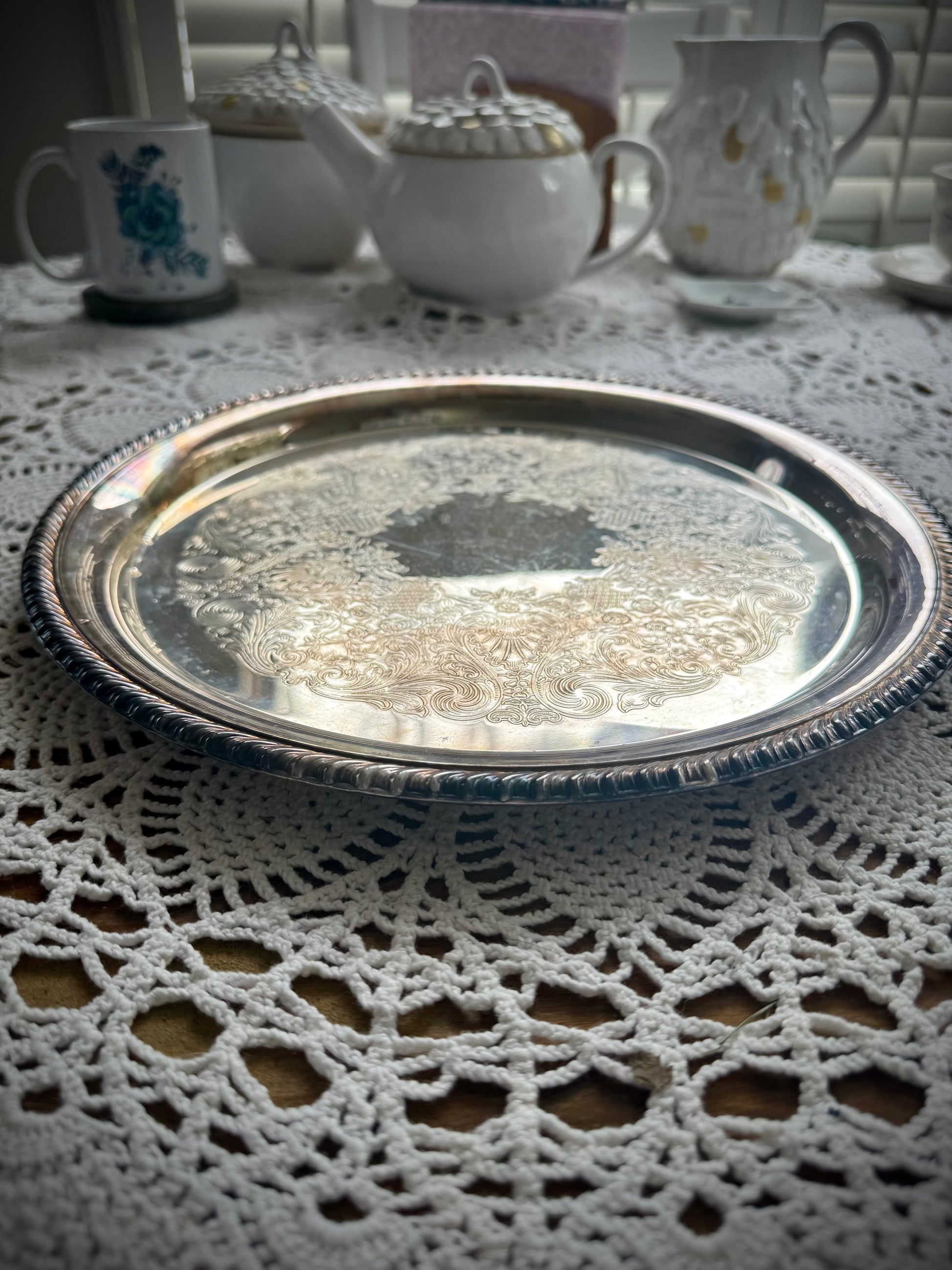 A silverplate round offering tray is displayed on a white lace surface. The tray is empty and has a reflective surface with ornate edge detailing.