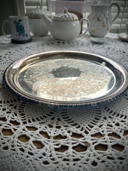 A silverplate round offering tray is displayed on a white lace surface. The tray is empty and has a reflective surface with ornate edge detailing.