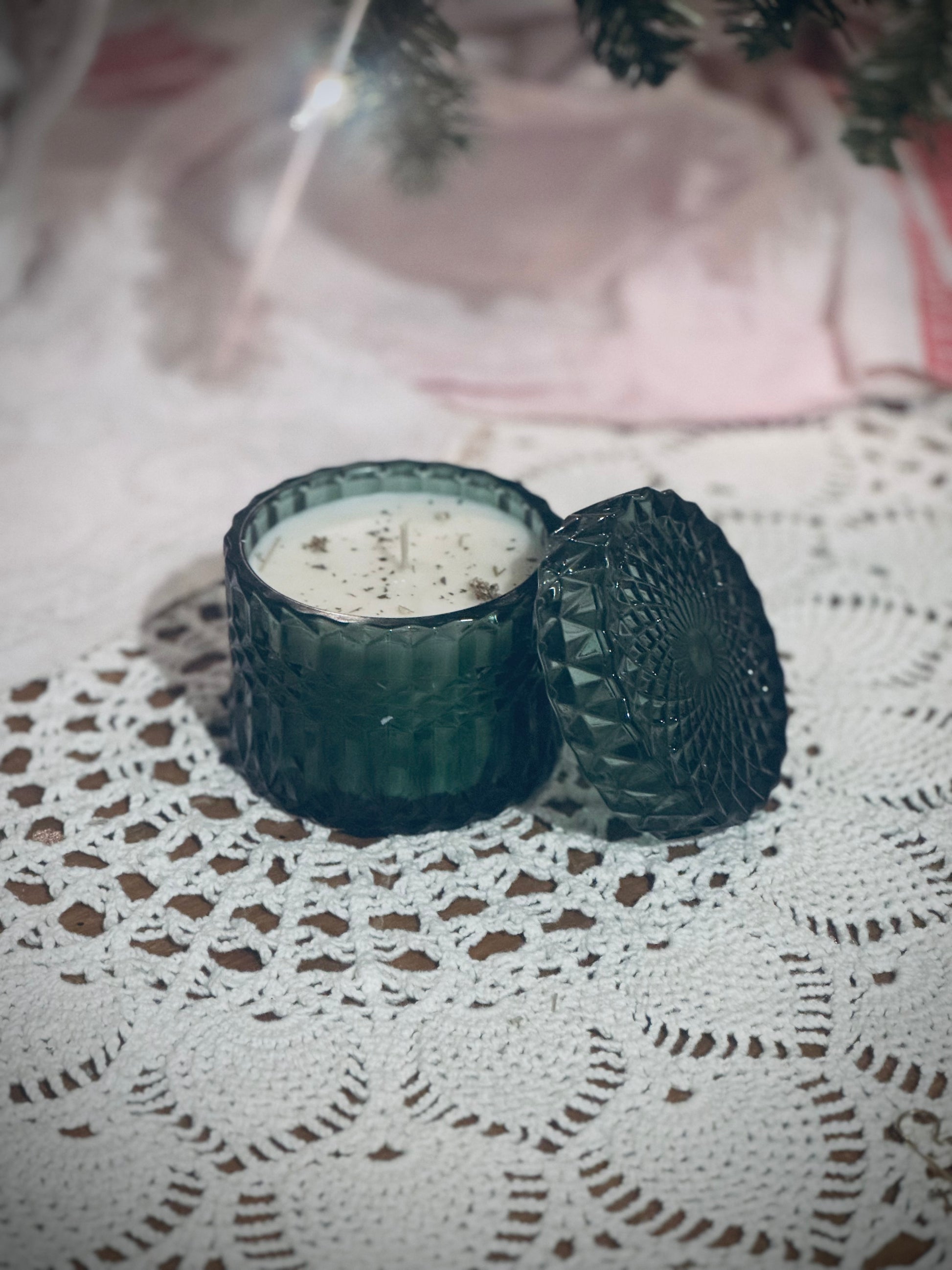 A green glass candle vessel with lid placed on a lace tablecloth in front of a decorated Christmas tree.