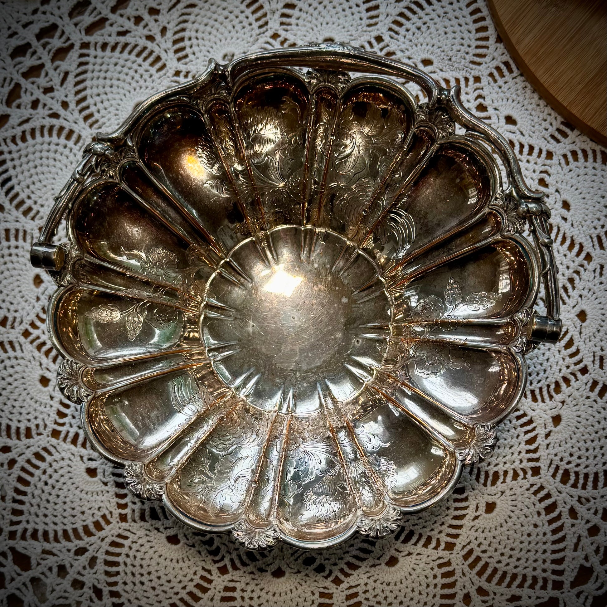 A silver plate basket with a handle, placed on a table with a lace tablecloth, displaying a reflective surface.