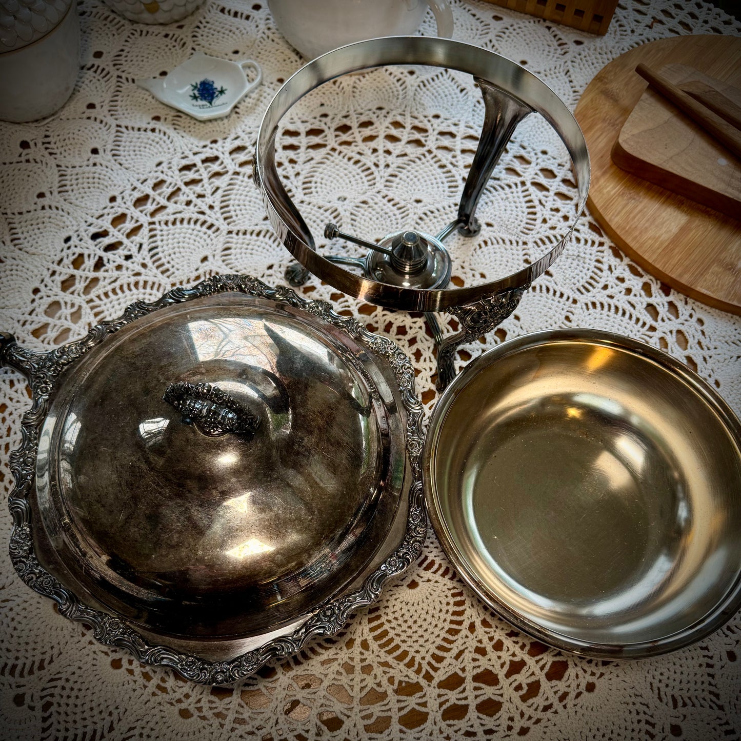 A vintage silver chafing dish with a lid, placed on a table with a lace tablecloth.