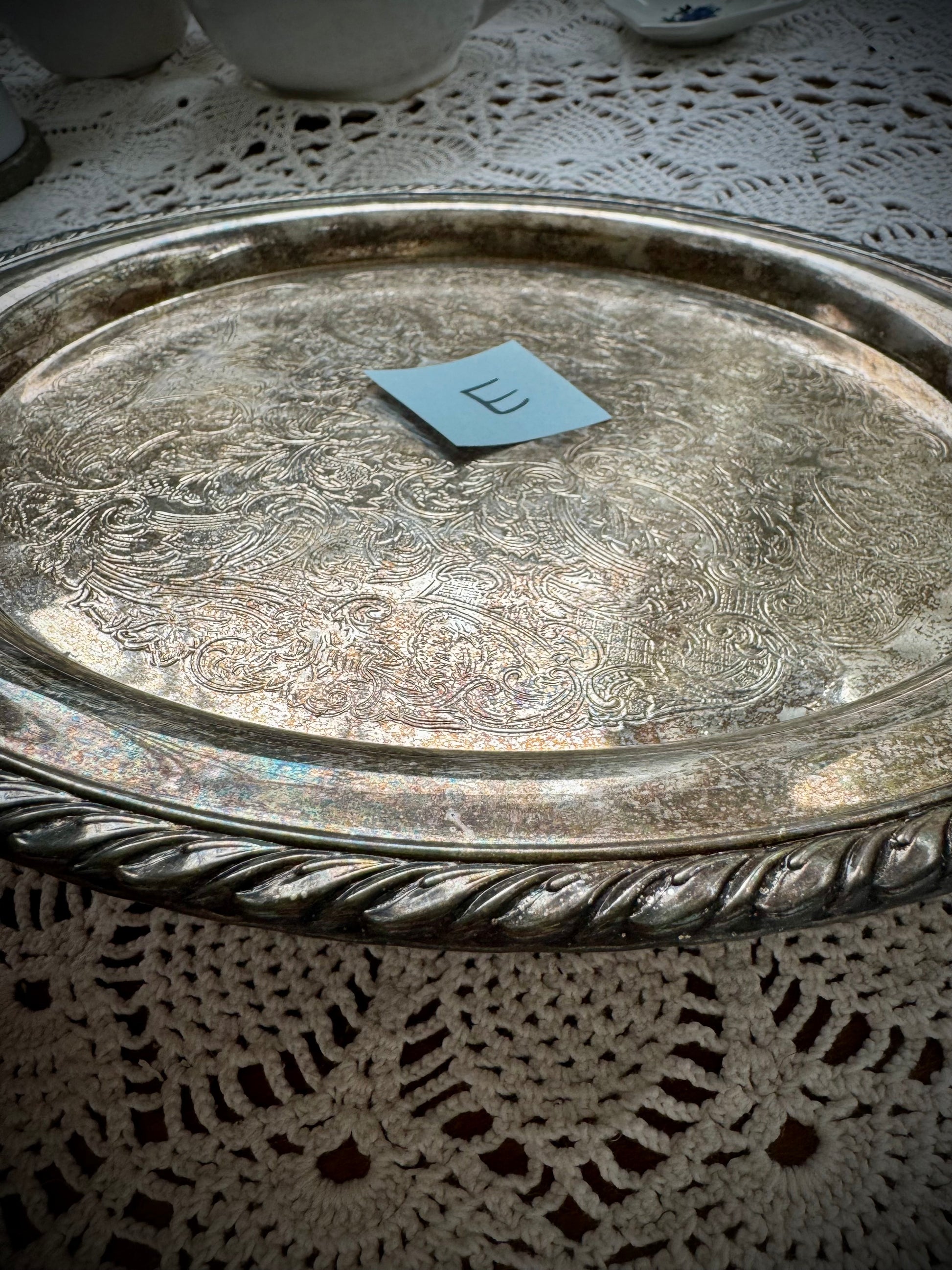 A silverplate round offering tray is displayed on a white lace surface. The tray has a post-it with the letter "E"