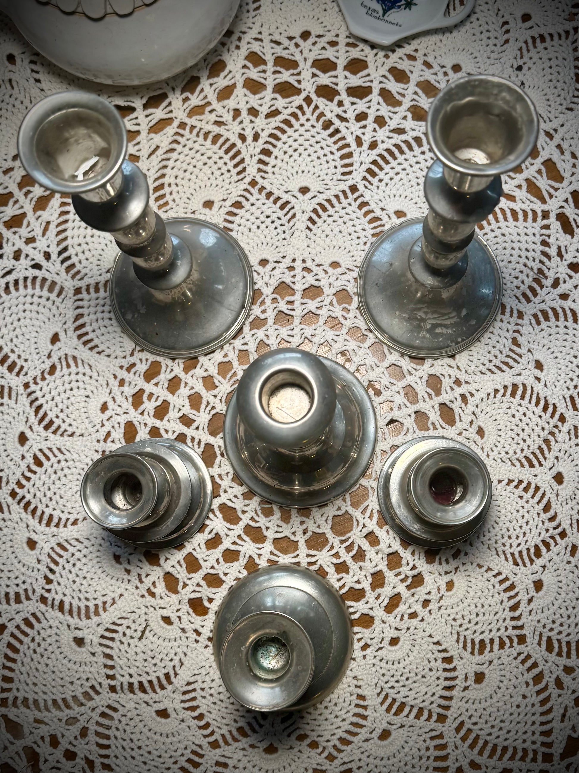 A collection of vintage pewter candlesticks in various sizes displayed on a table with a lace tablecloth.