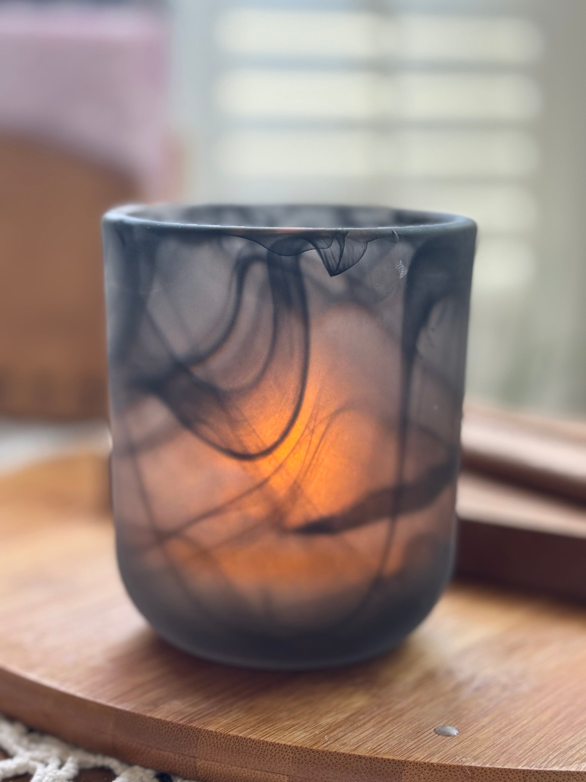 A black candle with black swirl patterns in a translucent vessel, sitting on a wooden surface with a warm light illuminated from the candle.