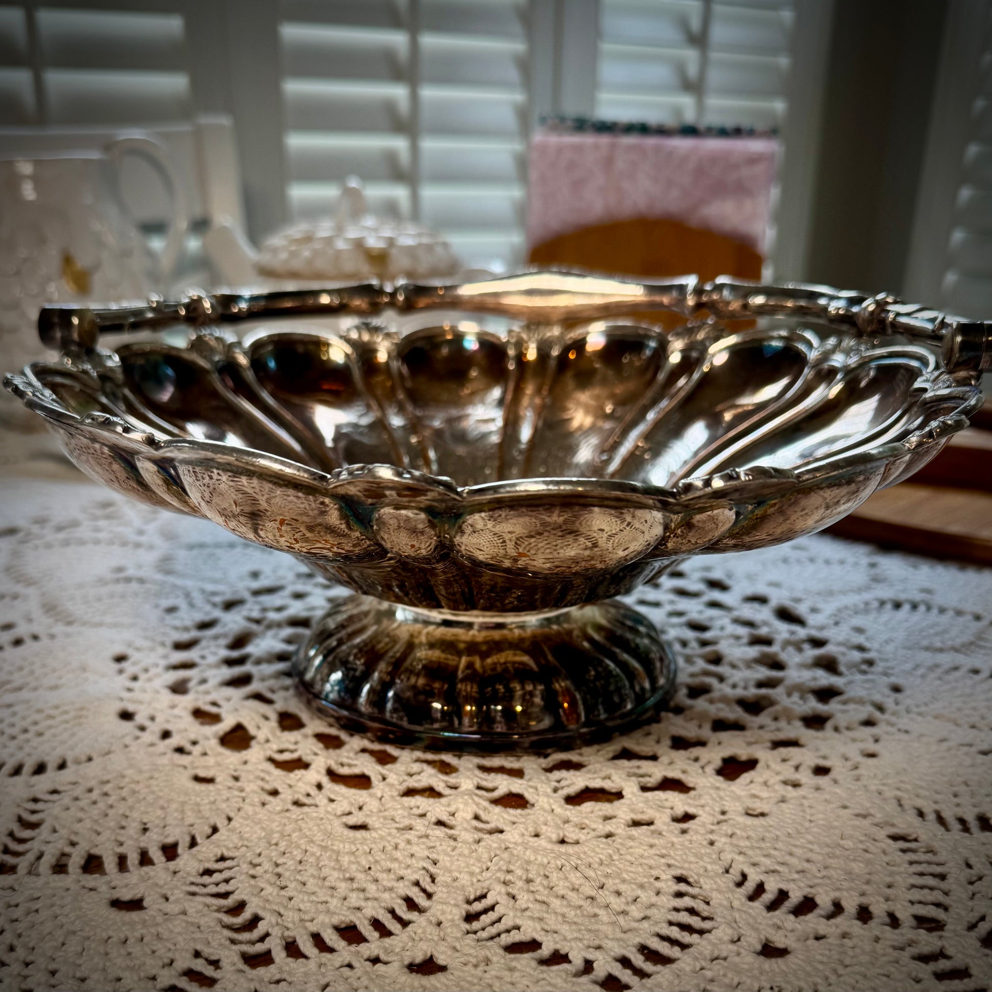 A silver plate basket with a handle, placed on a table with a lace tablecloth, displaying a reflective surface.