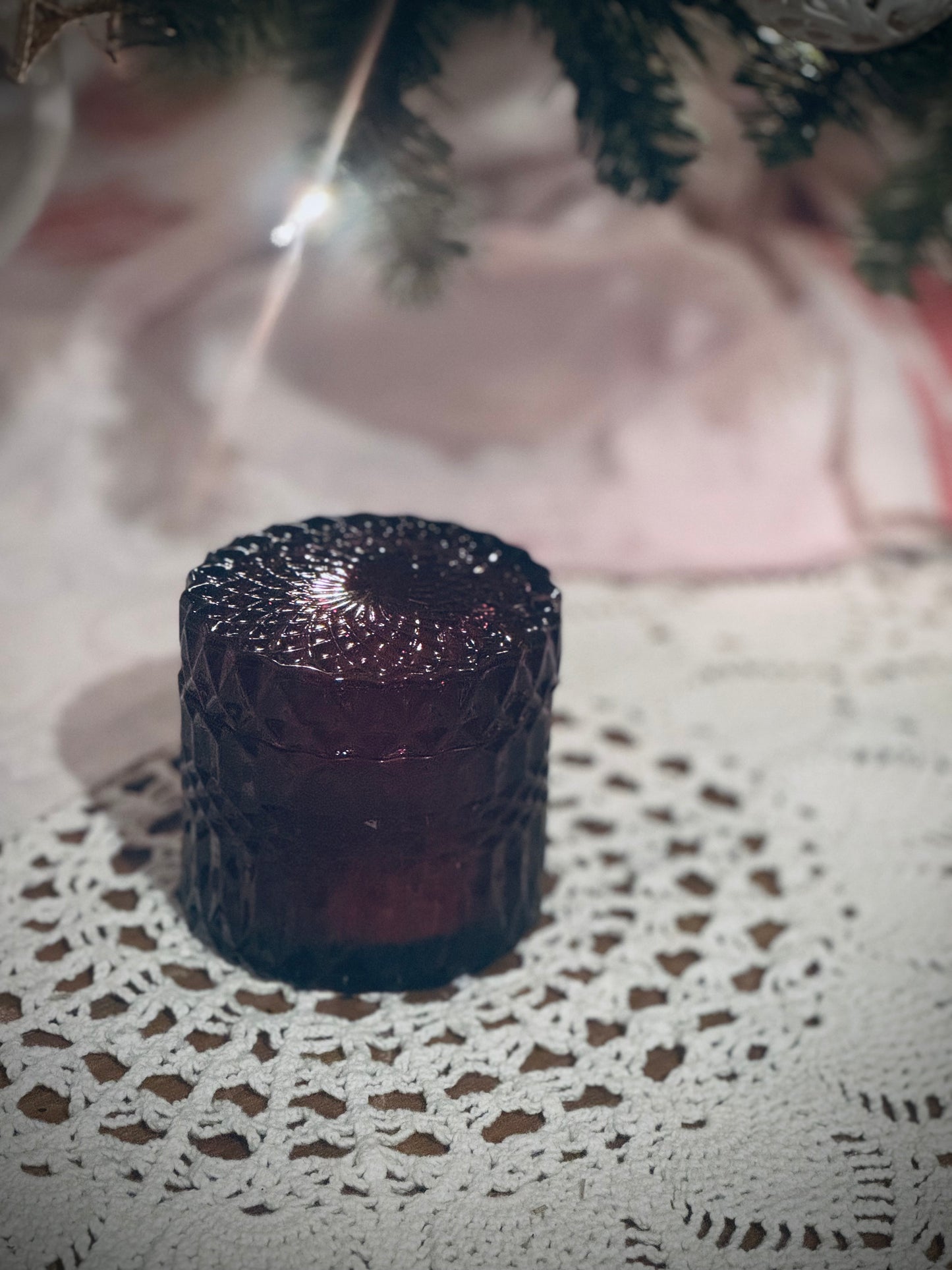 A deep red glass candle vessel with lid placed on a lace tablecloth in front of a decorated Christmas tree.