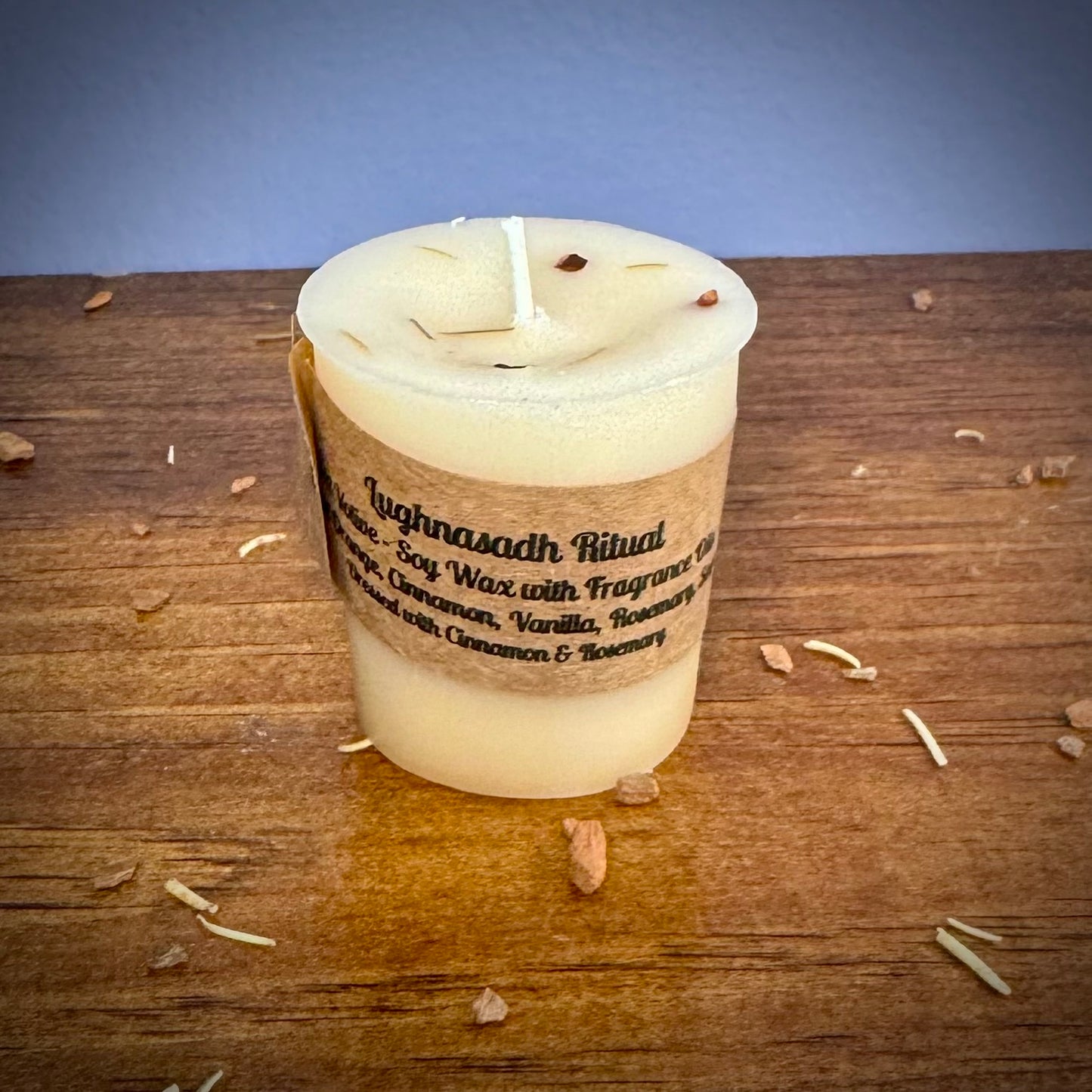 A white votive candle with a label reading "Lughnasadh Ritual" on a wooden surface.