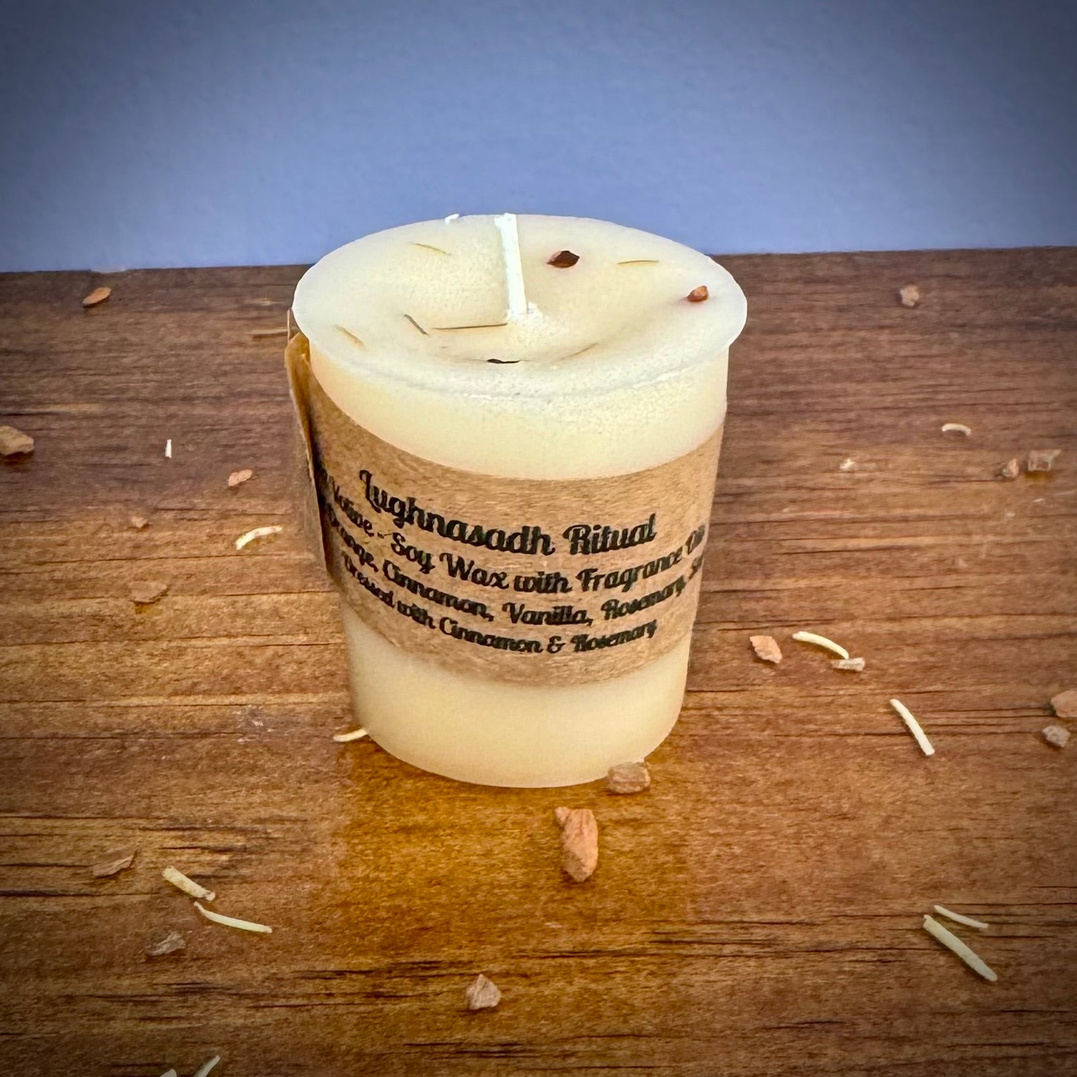 A white votive candle with a label reading "Lughnasadh Ritual" on a wooden surface.
