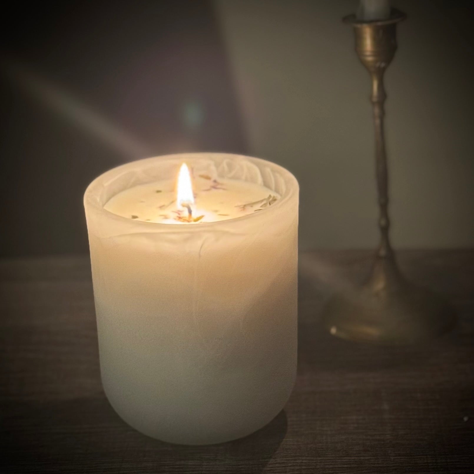 A white candle with a swirl vessel design, burning with a small flame, placed on a wooden surface with a metal candle holder in the background.