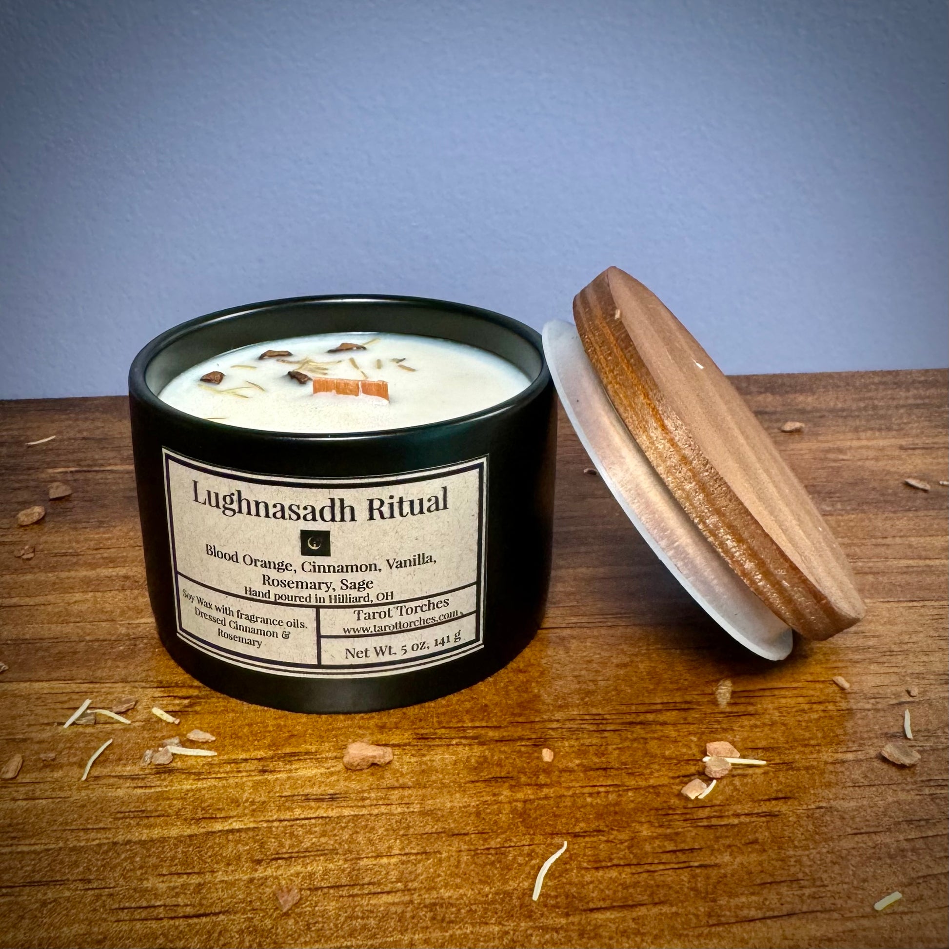 A ceramic candle labeled 'Lughnasadh Ritual' with an offset pine lid placed on a wooden surface against a purple background.