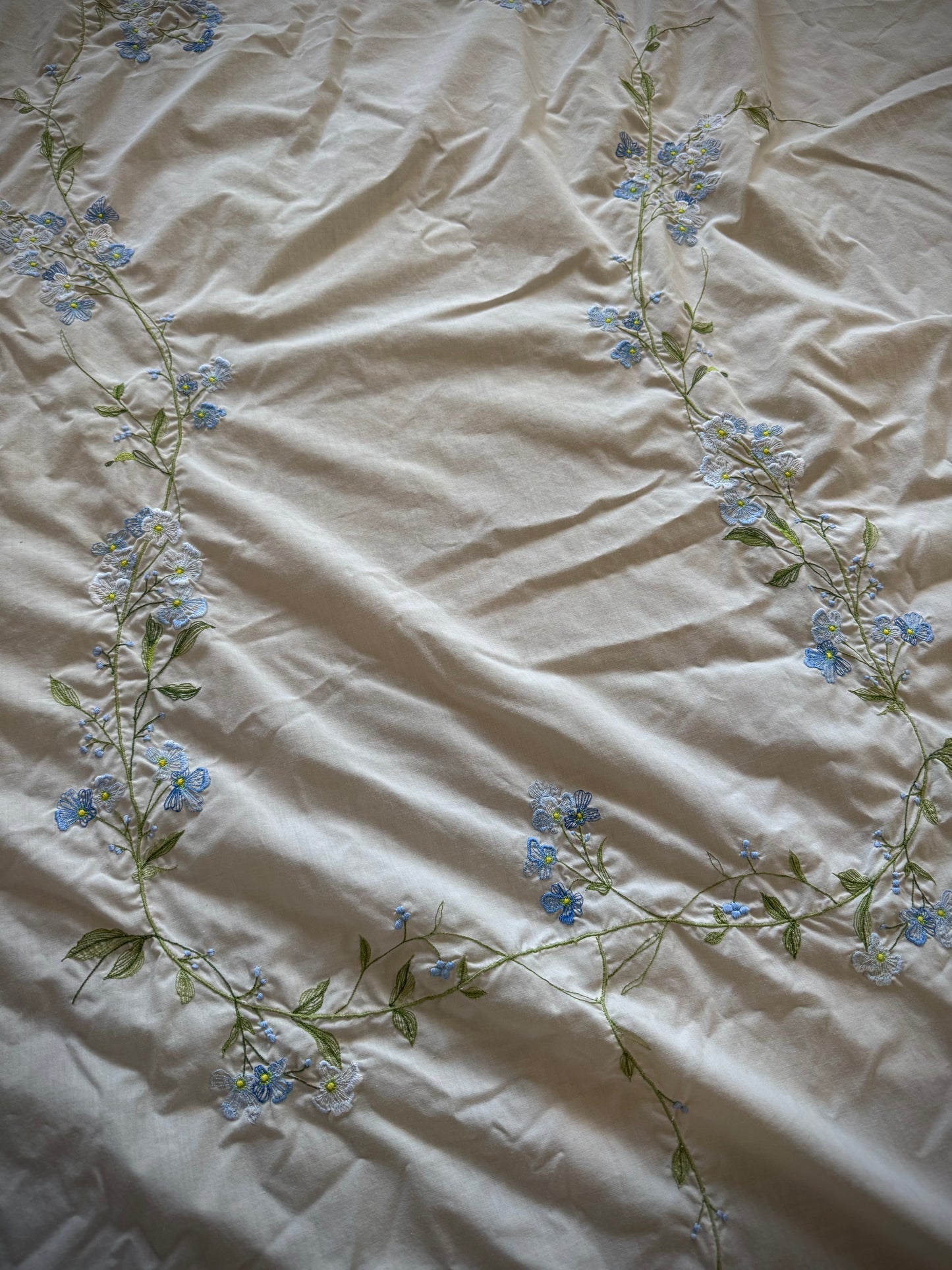 Vintage embroidered oval tablecloth laid out on a surface, primarily white with blue floral embroidery, showing signs of age-related discoloration.