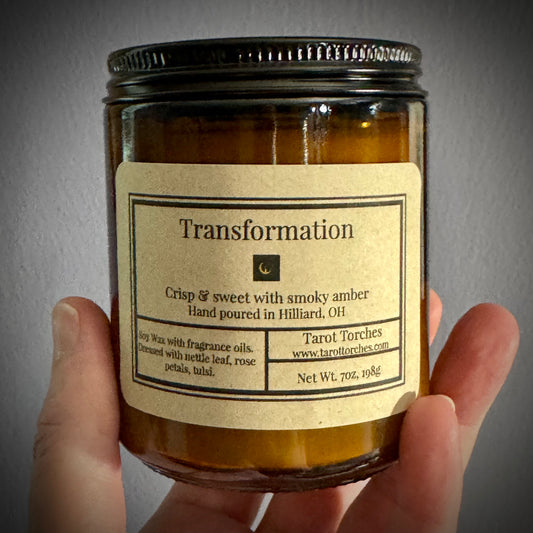 Candle labeled 'Transformation' held by a hand against a neutral background