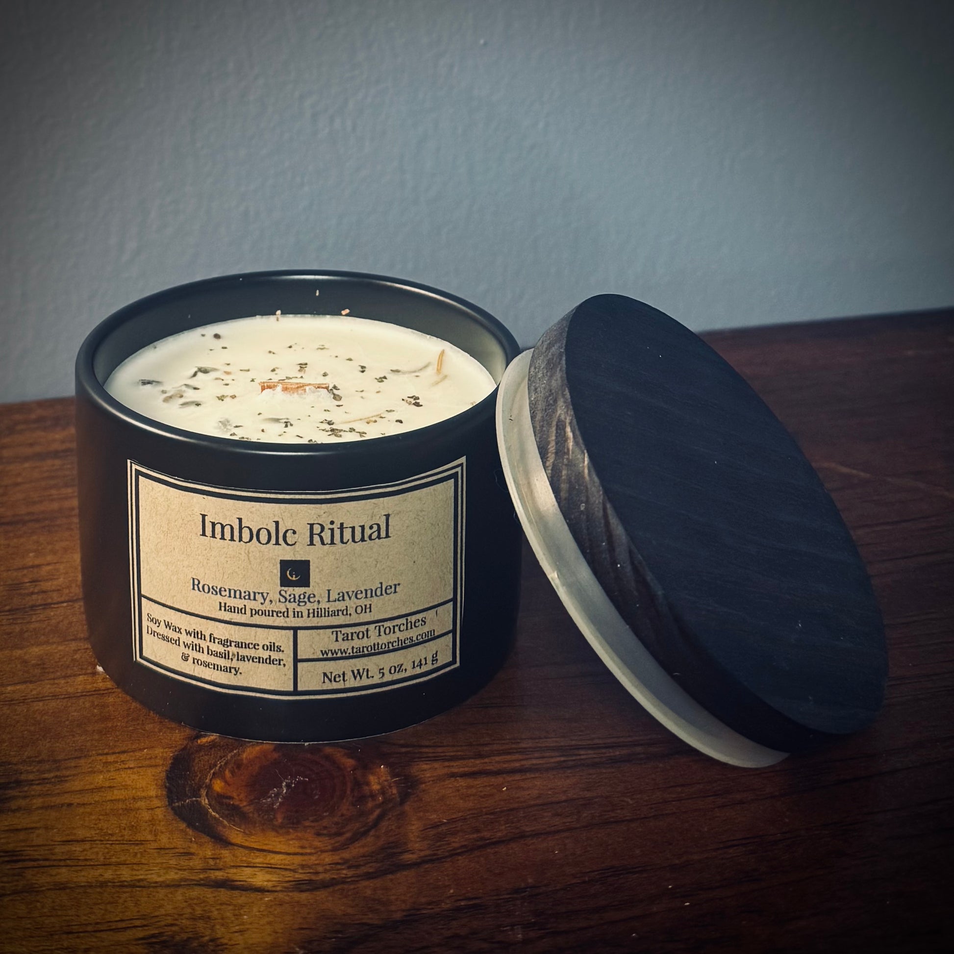A black ceramic candle with a lid, labeled 'Imbolc Ritual' with notes of rosemary, sage, and lavender, sitting on a wooden surface.