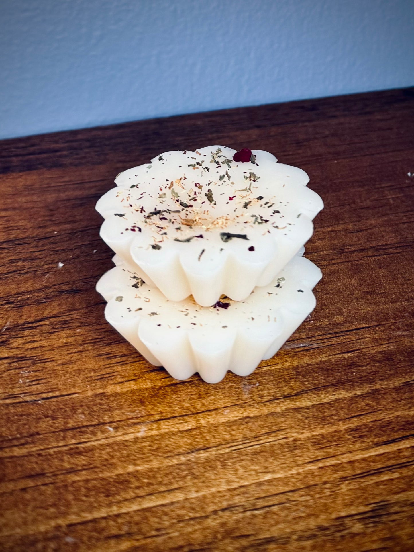 A stack of two white soy wax tarts with visible herbs on a wooden surface.